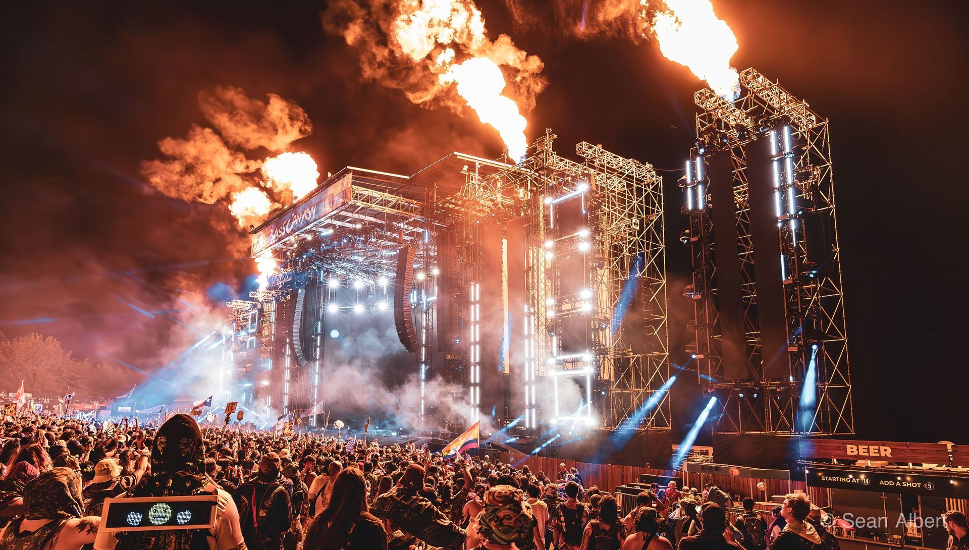 Concert stage at night with flames, fireworks, and large crowd of people.