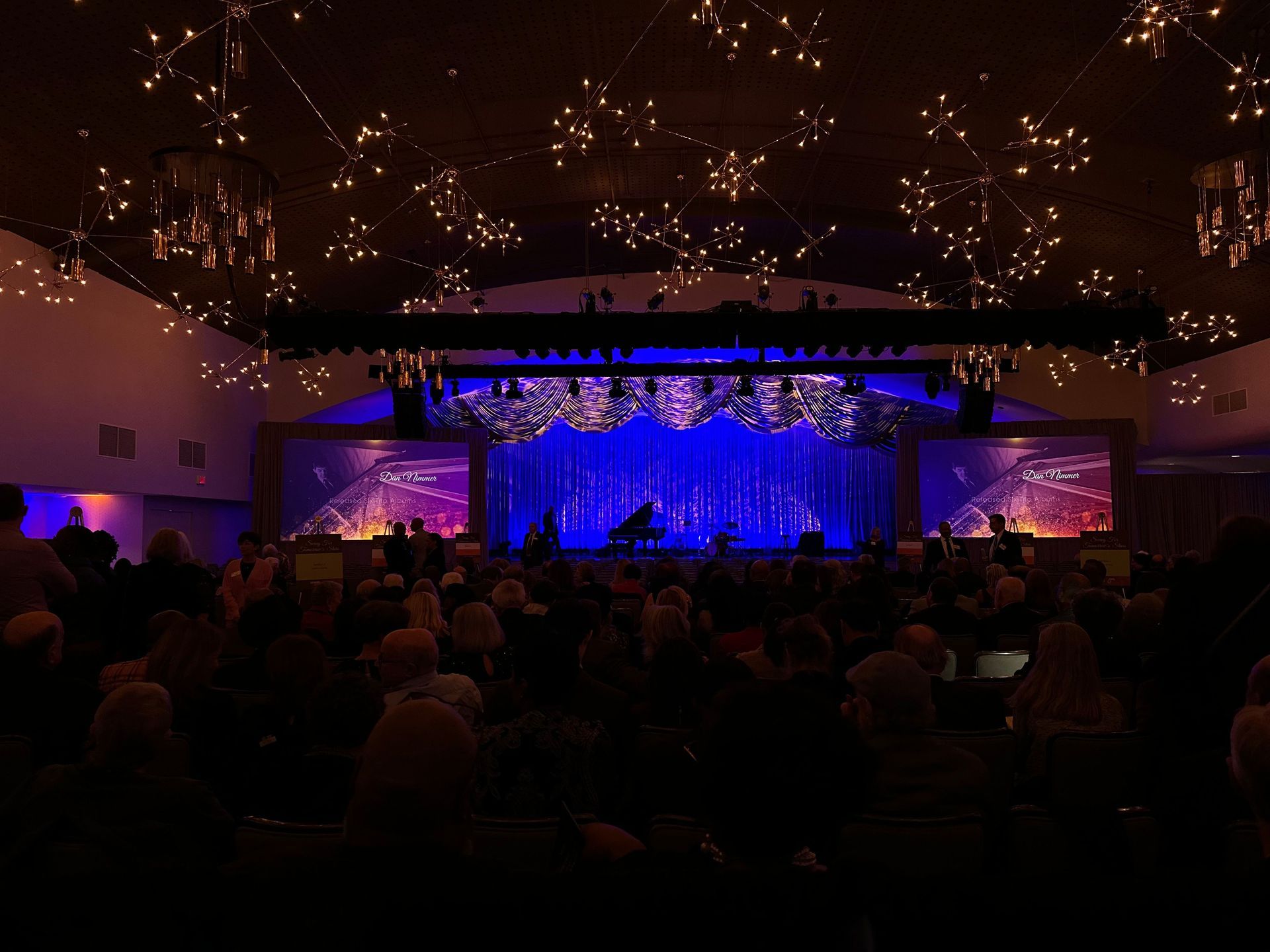 Audience in a dark auditorium facing a stage with blue and purple lighting, and star-shaped lights on the ceiling.