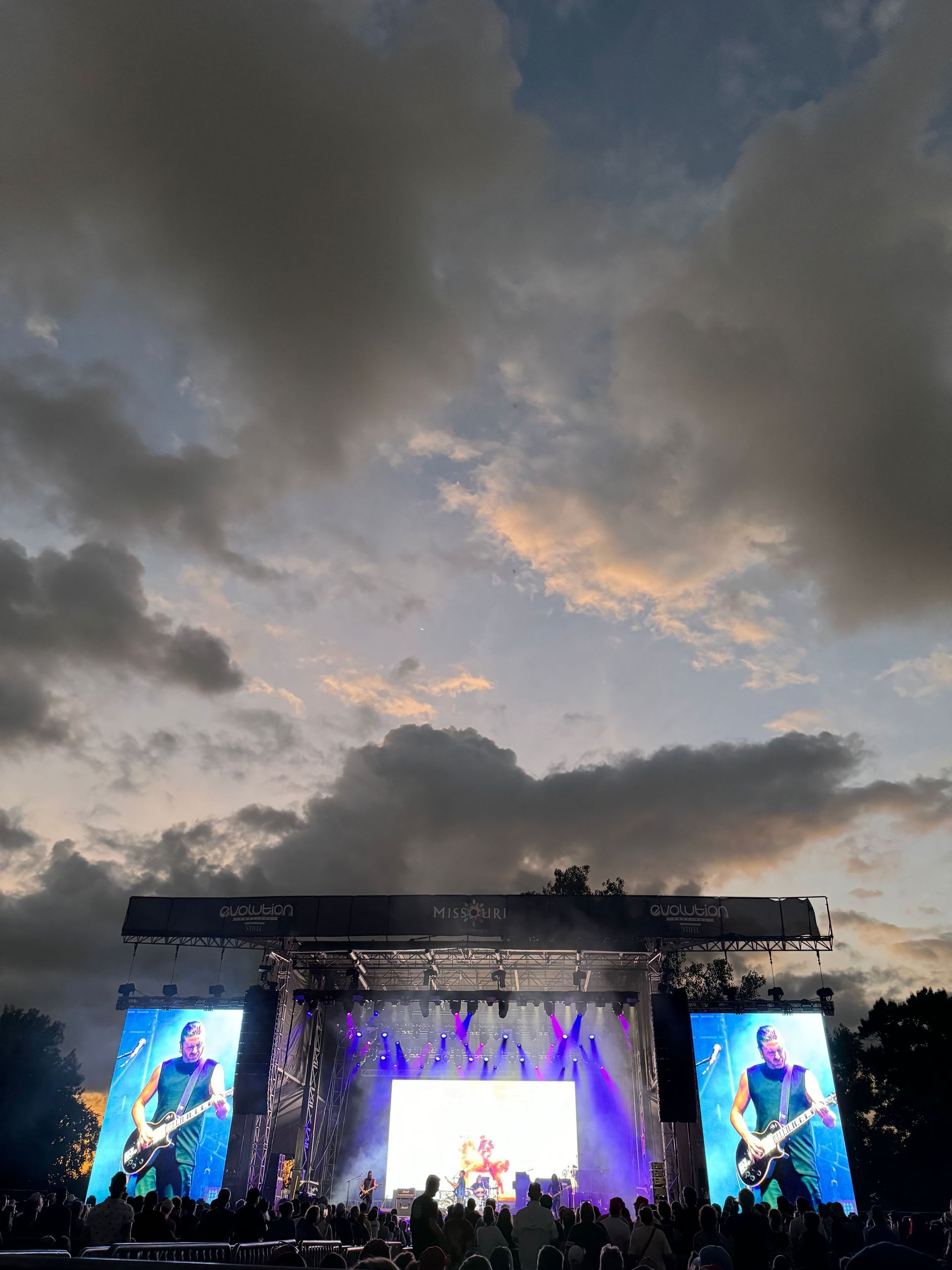 Concert stage under a cloudy sky. Large screens show performers. Audience in silhouette. Dusk setting.