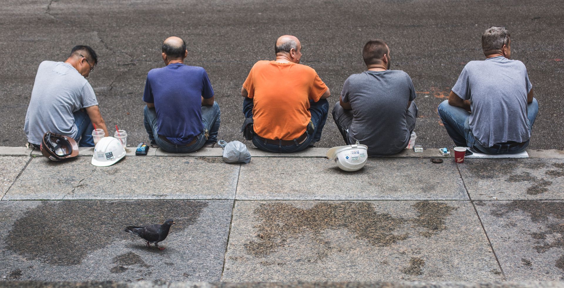 A group of men are sitting on the sidewalk looking at a pigeon.