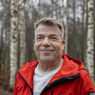 A man in a red jacket is smiling in front of a forest.