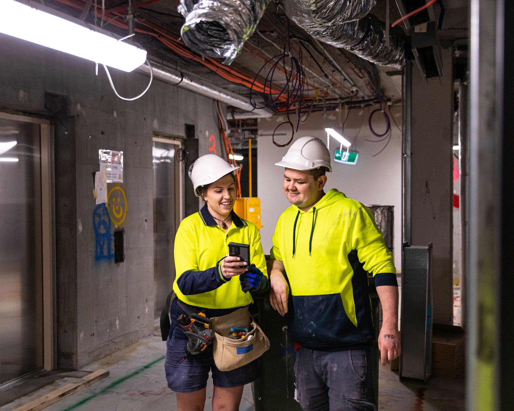 A man and a woman are standing in a hallway looking at a cell phone.