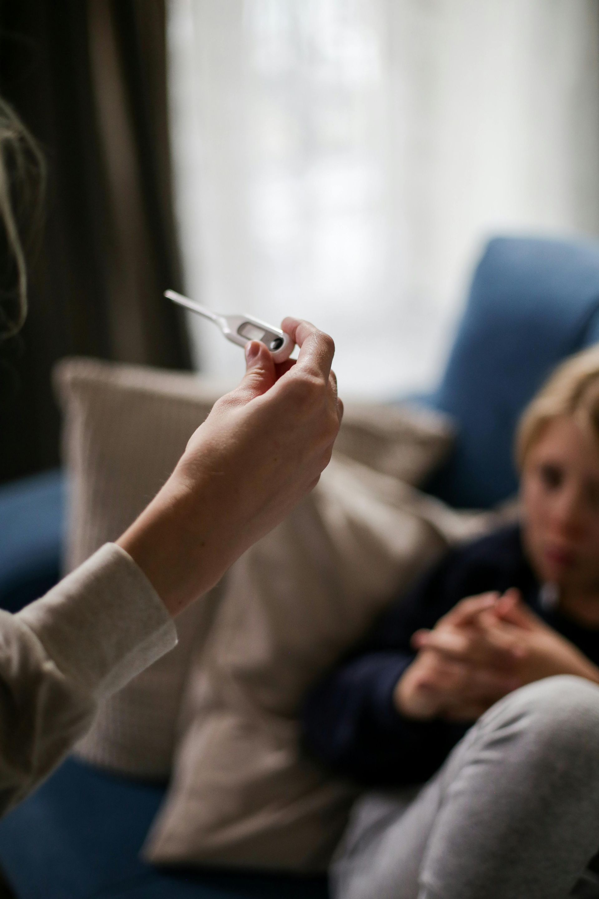 Person som håller termometer och mäter temperaturen på en person som sitter i en blå soffa.