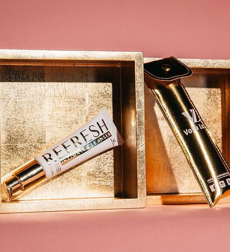 Two cosmetic tubes in gold display boxes on a pink background, one labeled “REFRESH.”