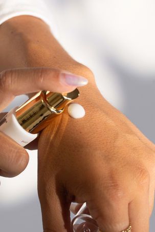 Hand dispensing white cream onto the back of a hand with a gold-topped pump bottle