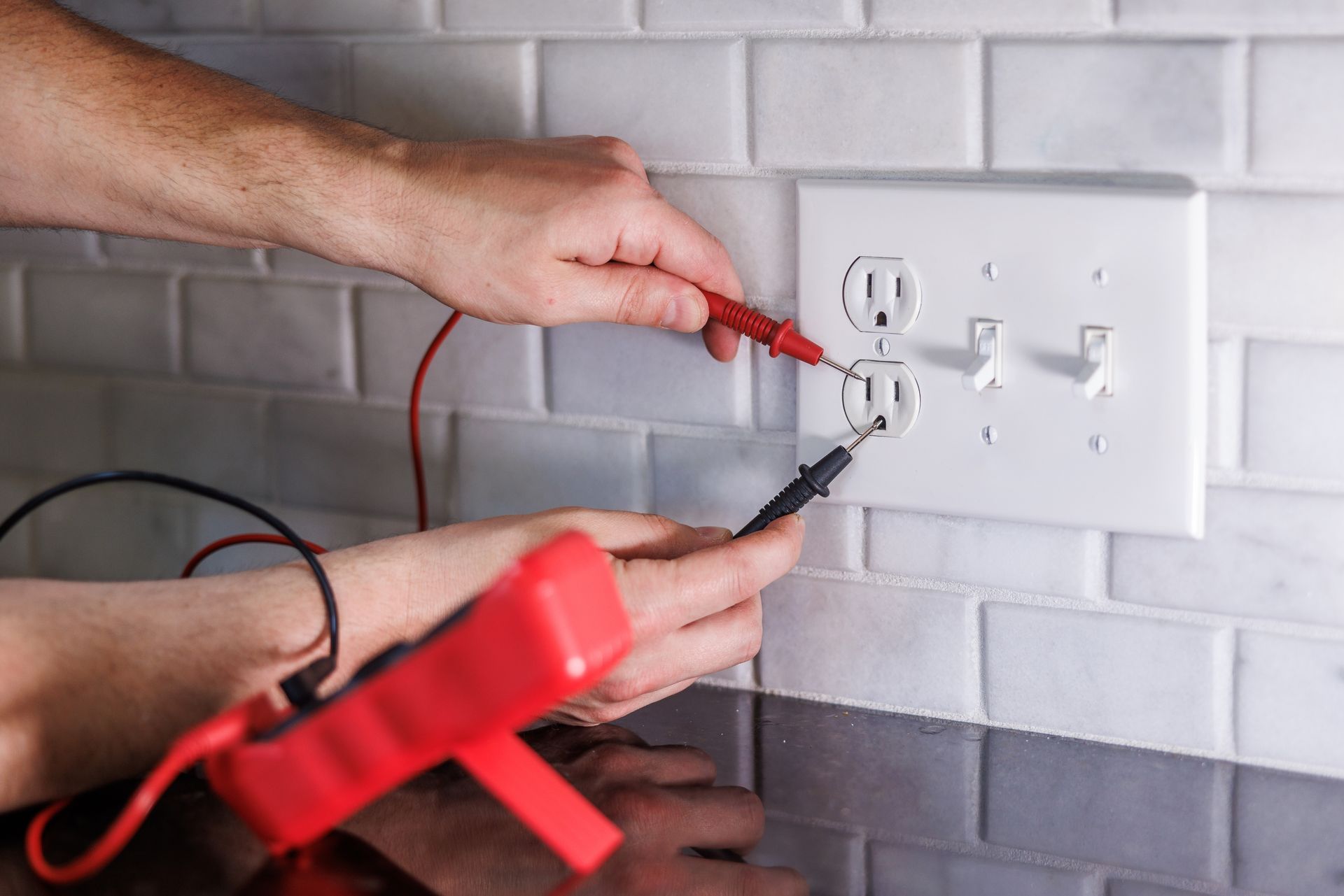Hands testing an electrical outlet with a multimeter, on a tiled wall.