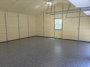 An empty garage with a grey speckled epoxy floor, insulated beige walls, a small window, and overhead door tracks.