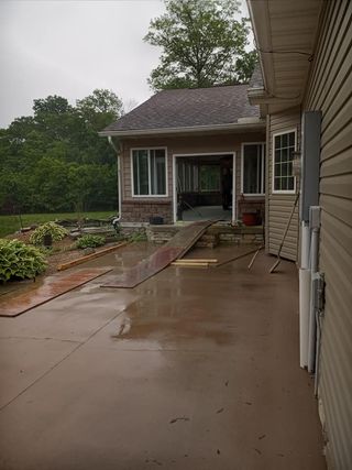 A wet concrete patio leads to a doorway with a makeshift ramp leading into an open room of a tan-sided house.