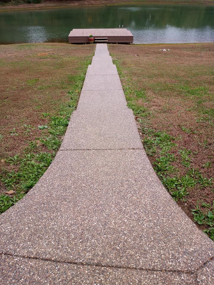 A long, straight concrete path leading down through a grassy yard to a rectangular dock on a calm lake.