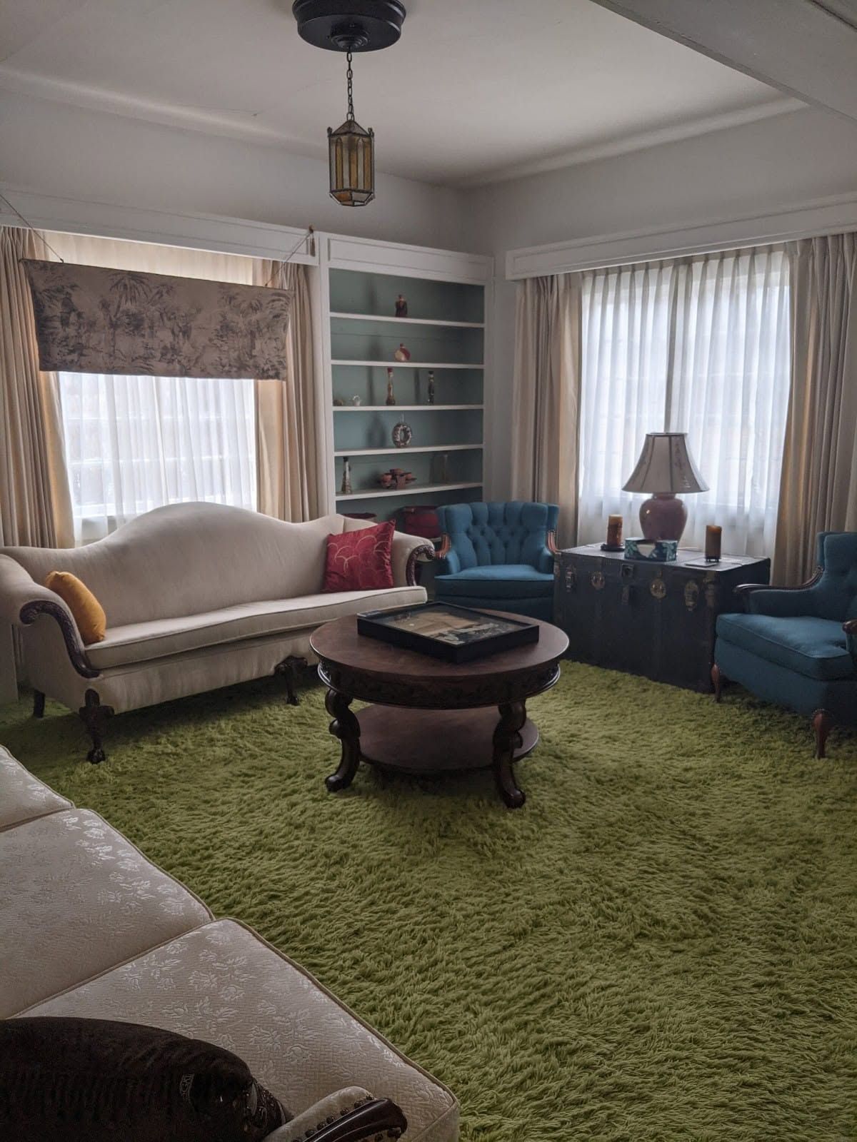 Bright living room with green shag carpet, sofas, a round coffee table, and large windows with curtains