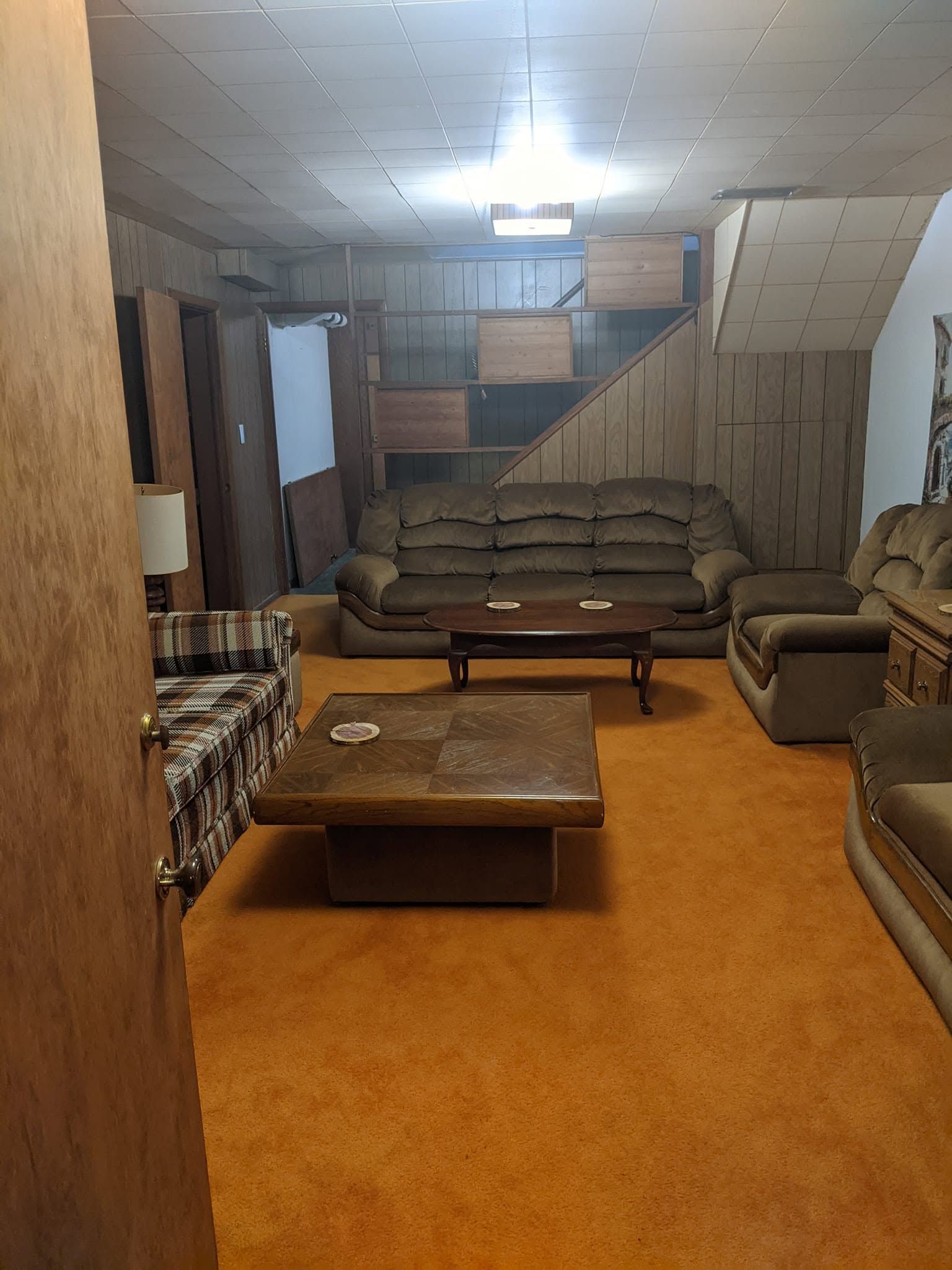 Dim basement living room with orange carpet, brown sofa, recliners, coffee table, and stairs at the back.