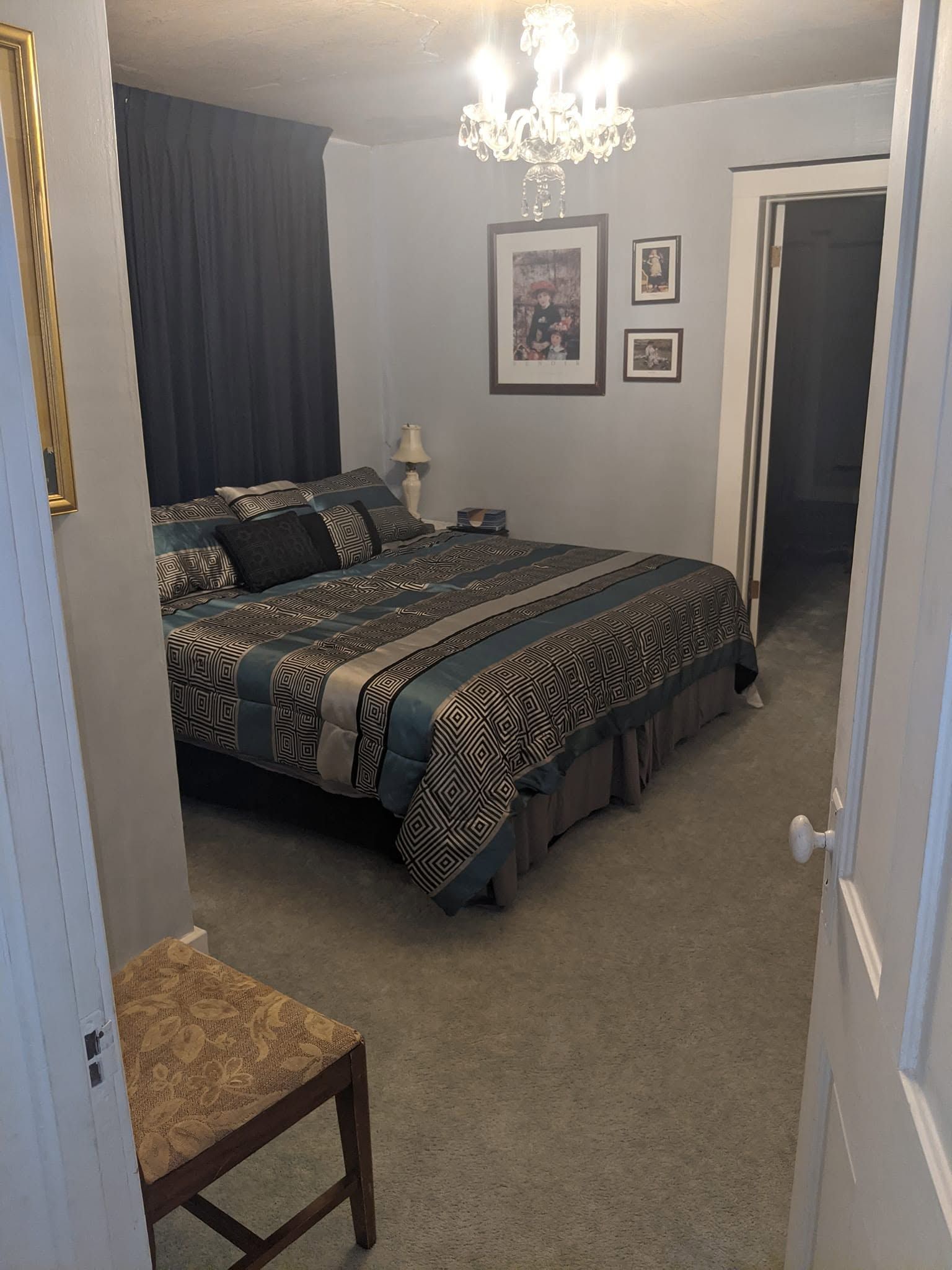 Bedroom with a bed, dark curtains, chandelier, framed art, and an open doorway to a hall.