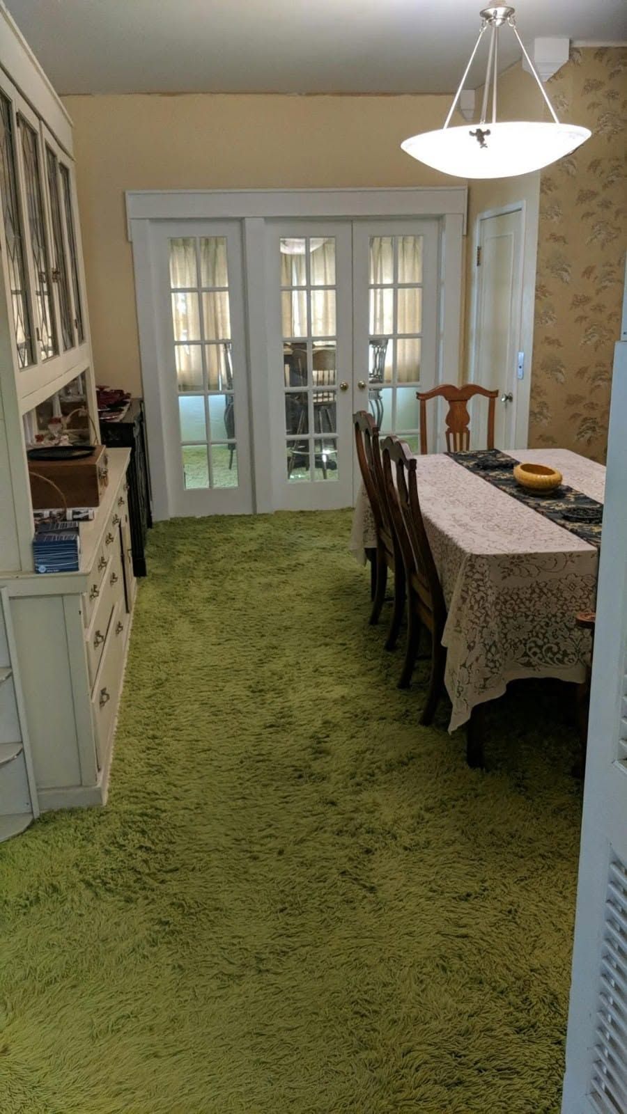 Dining room with a long table and chairs on green carpet, lit by a ceiling pendant and French doors at the back.