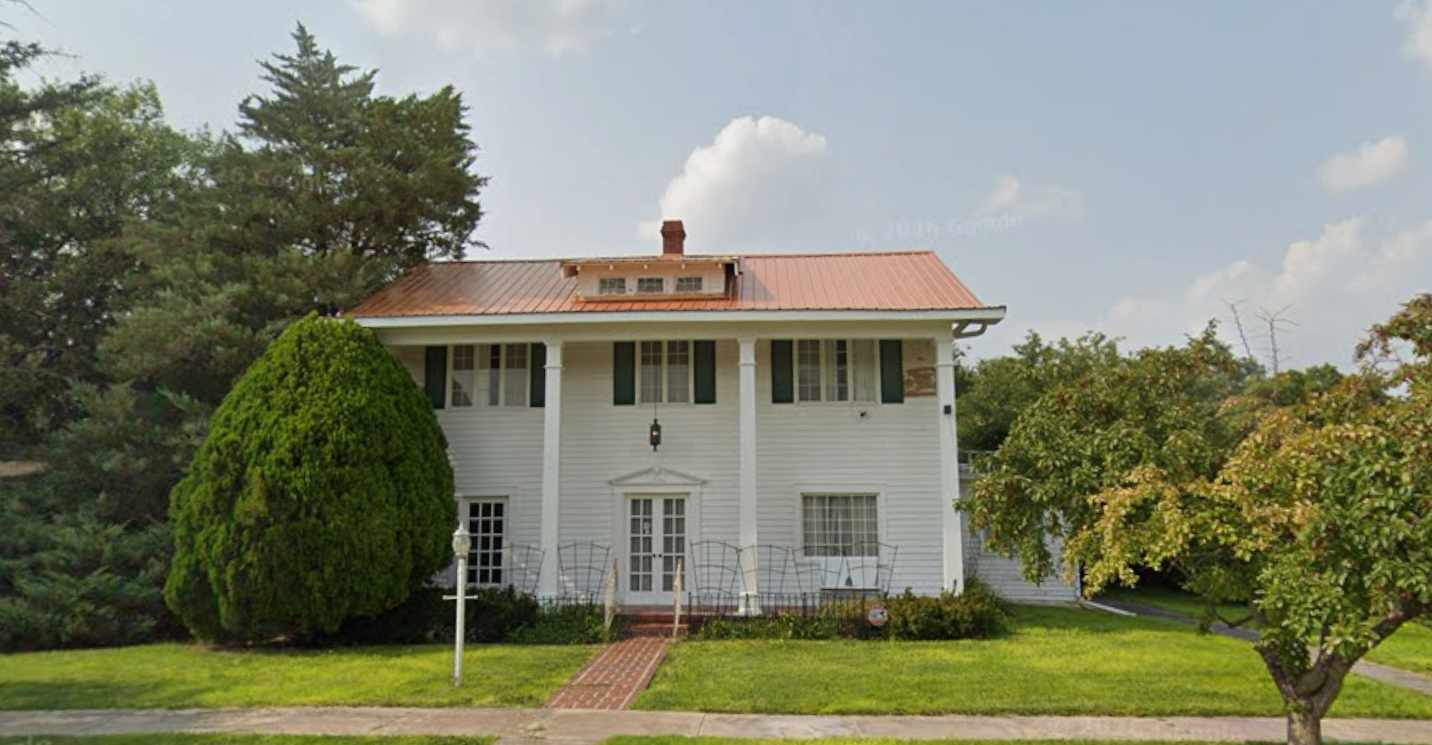 White two-story house with red roof, centered behind a brick path and green lawn, surrounded by trees