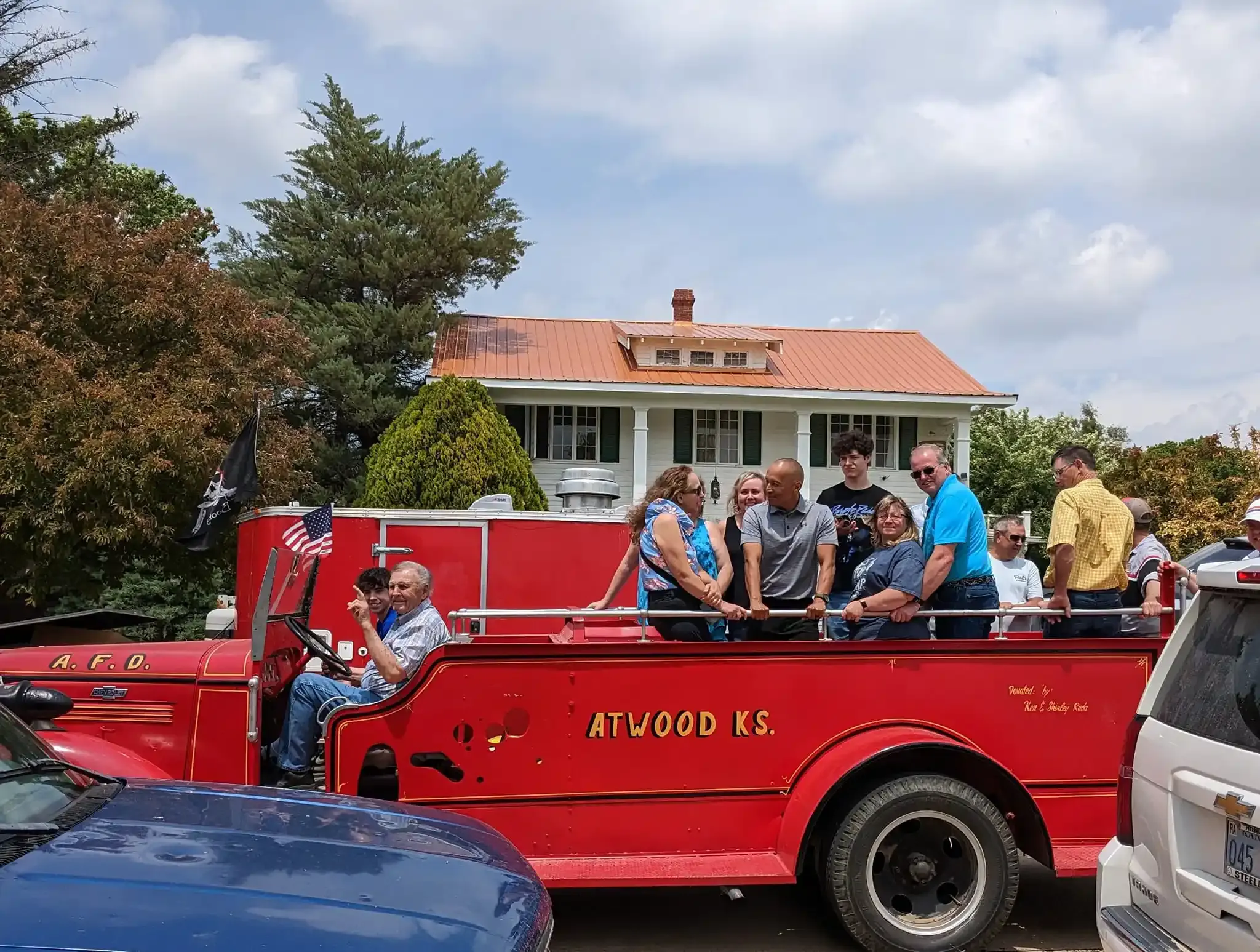 People riding in a red open-top truck on a street, with houses and trees in the background.
