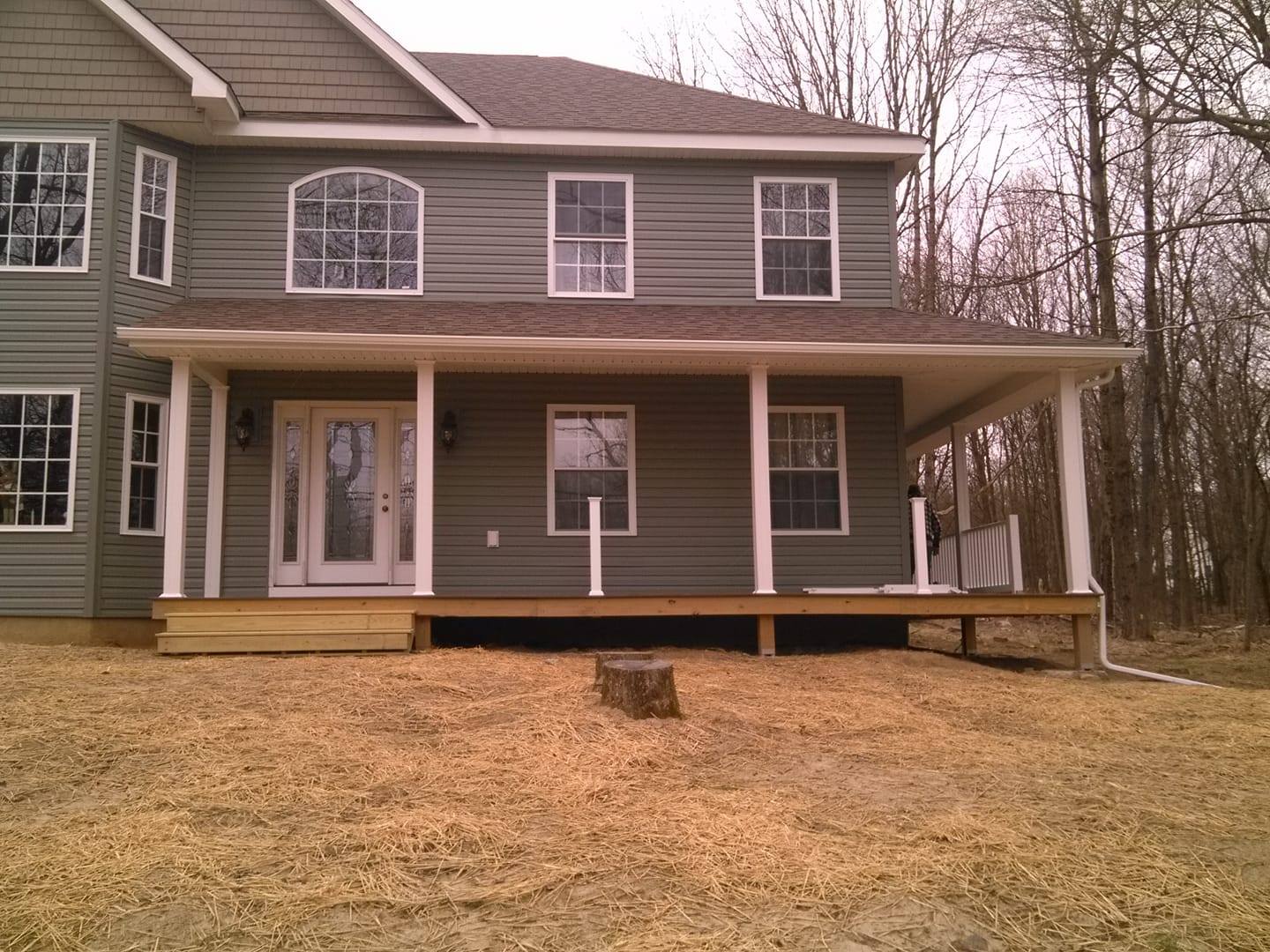 Two-story green house with a covered front porch and wooden deck on a grassy lot.
