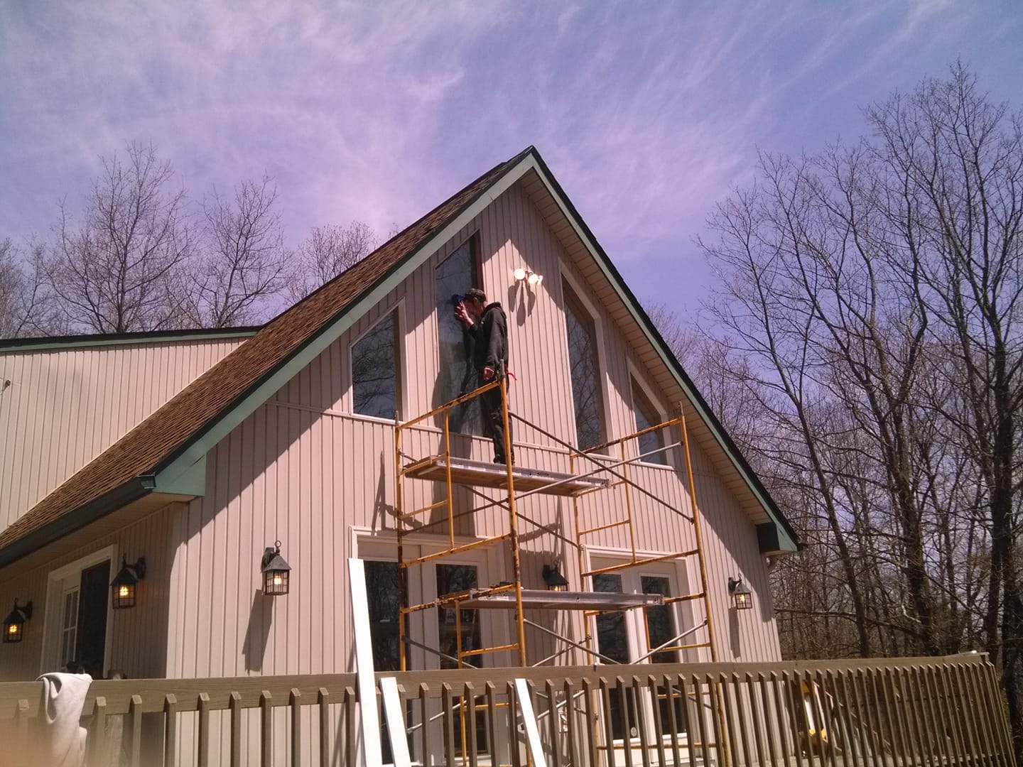 Worker on scaffolding painting the front gable of a beige house with a wooden fence and bare trees nearby.