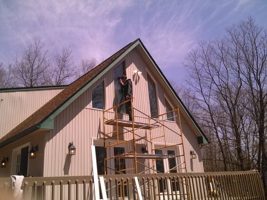 Worker on scaffolding painting the front gable of a beige house with a wooden fence and bare trees nearby.