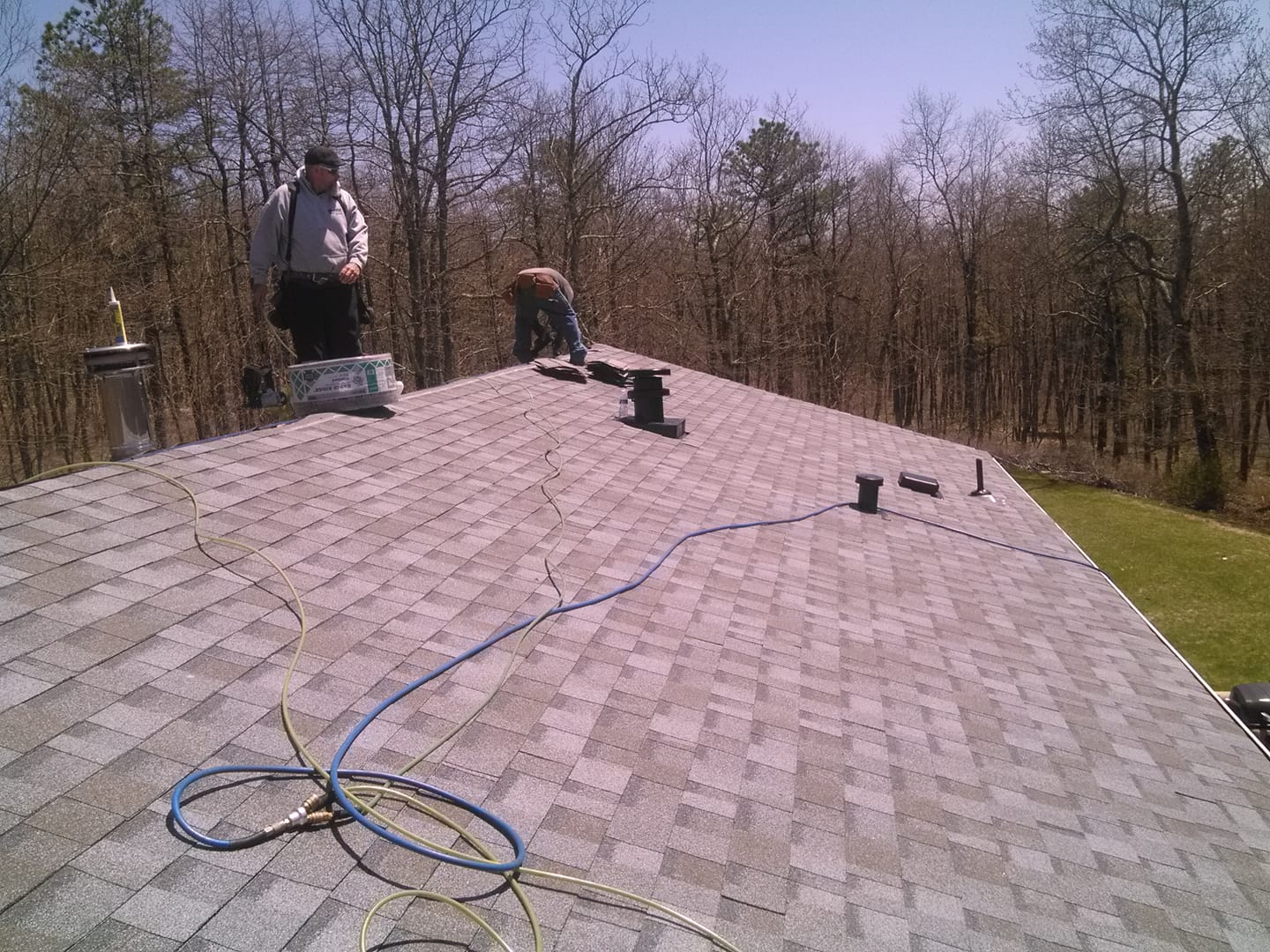 Workers on a shingled roof with tools and coiled hose, surrounded by trees on a sunny day