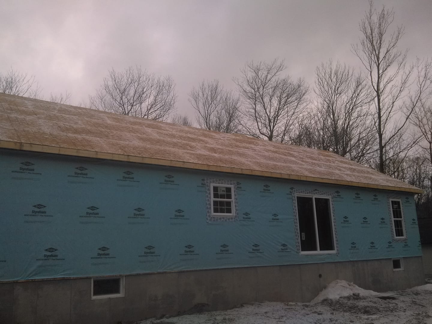 Partially built house with blue house wrap, roof, and leafless trees under a cloudy sky