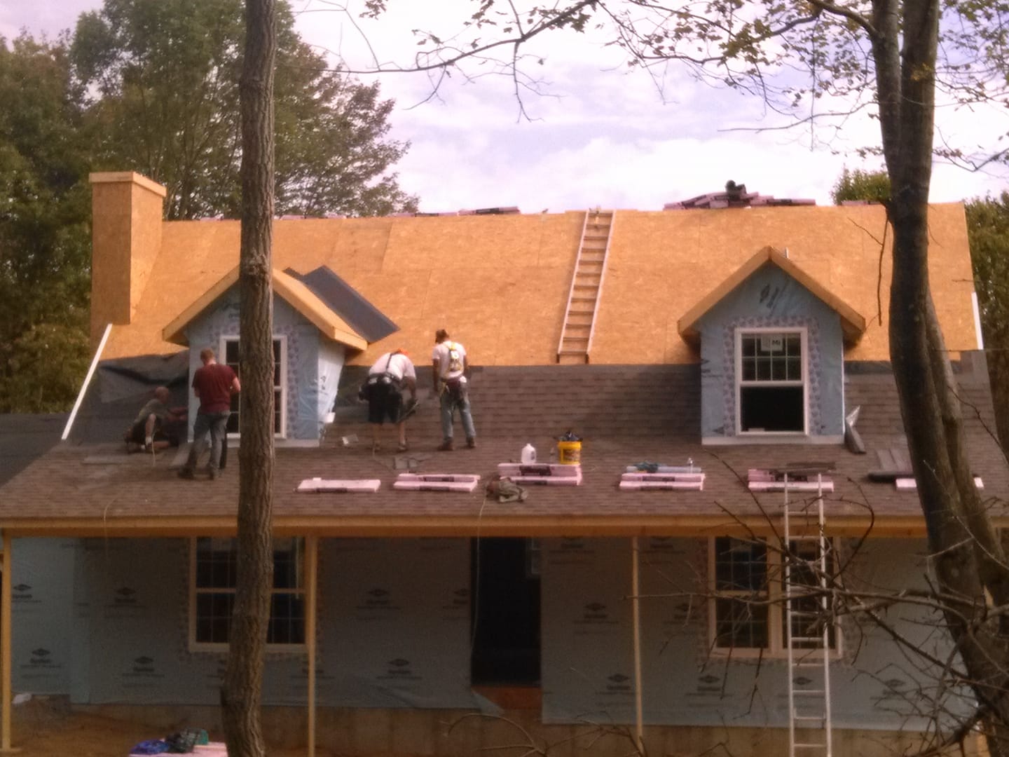 Workers installing a tan roof on a house under construction, with ladders and tools visible.