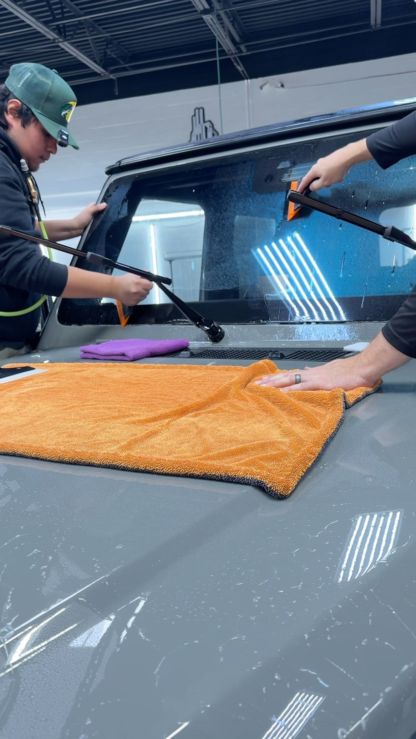 Two workers wiping a car windshield with cloths, one orange, one black.