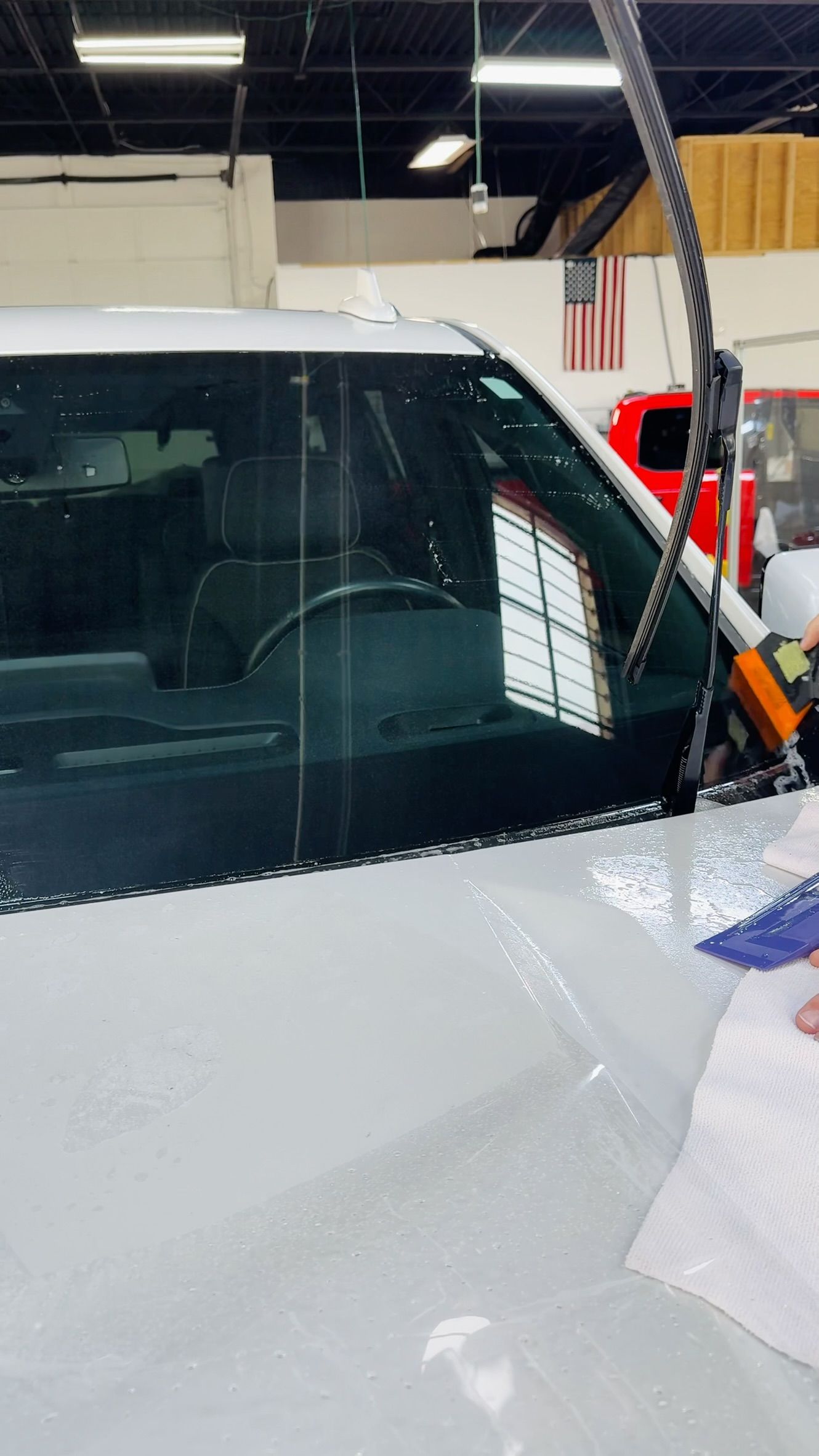 White car windshield being cleaned in an auto shop, with a long tool and cloth visible.