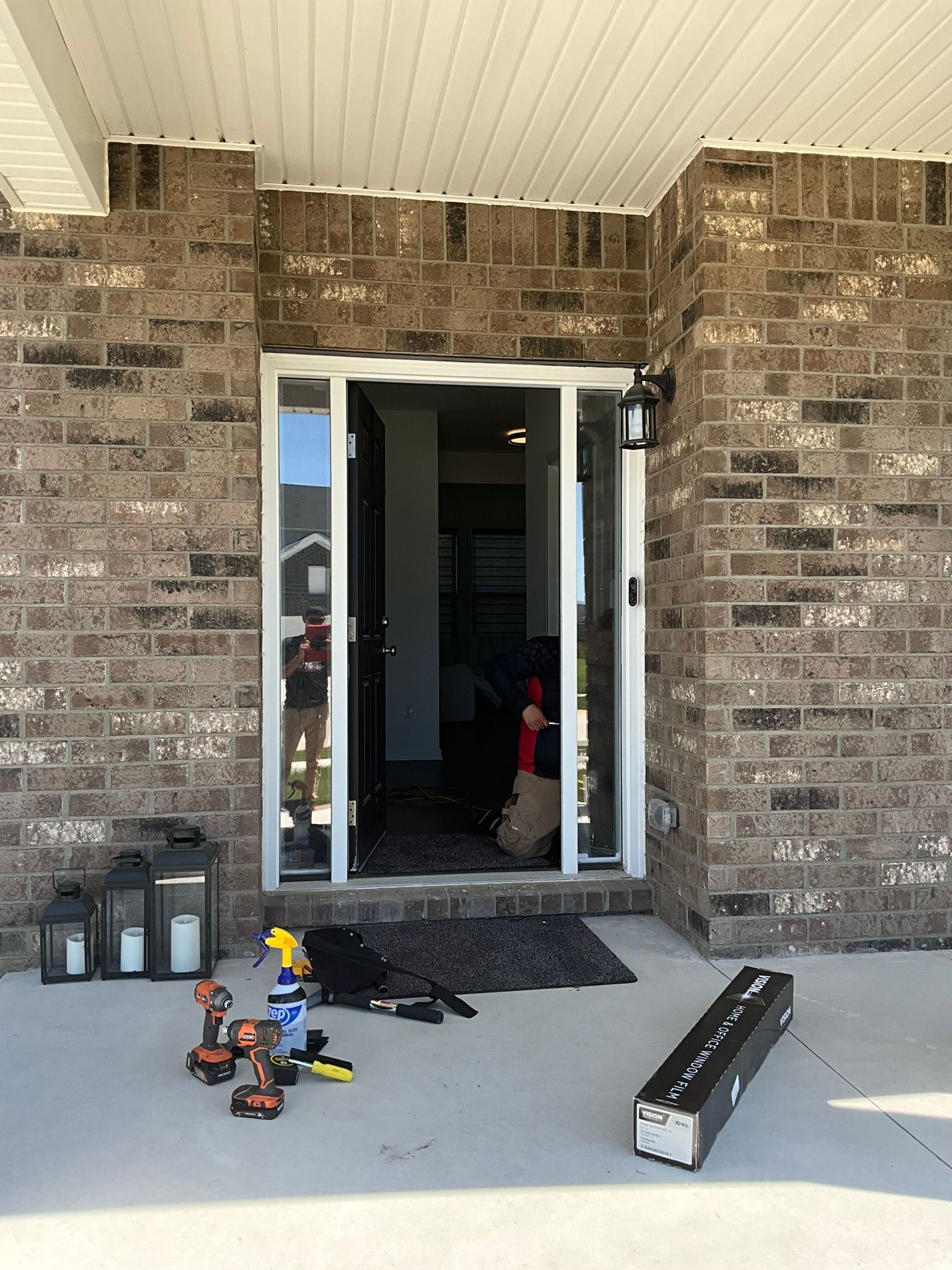 Front porch with open glass door, brick facade, lanterns, doormat, and scattered toys on the concrete floor.