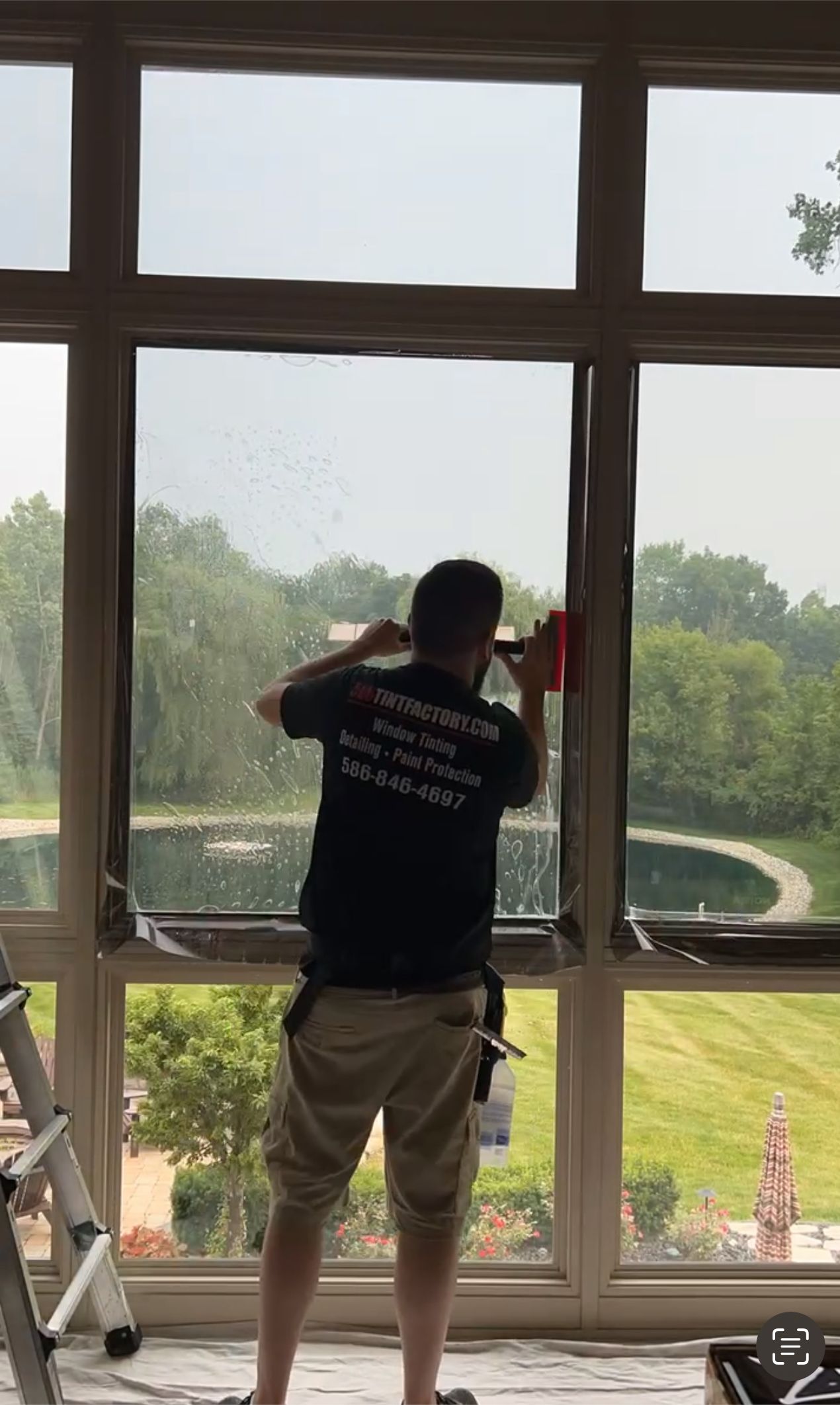 Person standing by large window, looking out at a green yard and trees.