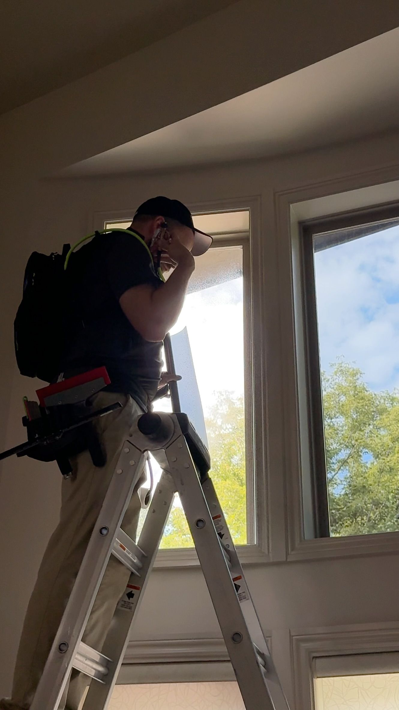 Worker on a ladder installing or inspecting a ceiling light near a bright window.