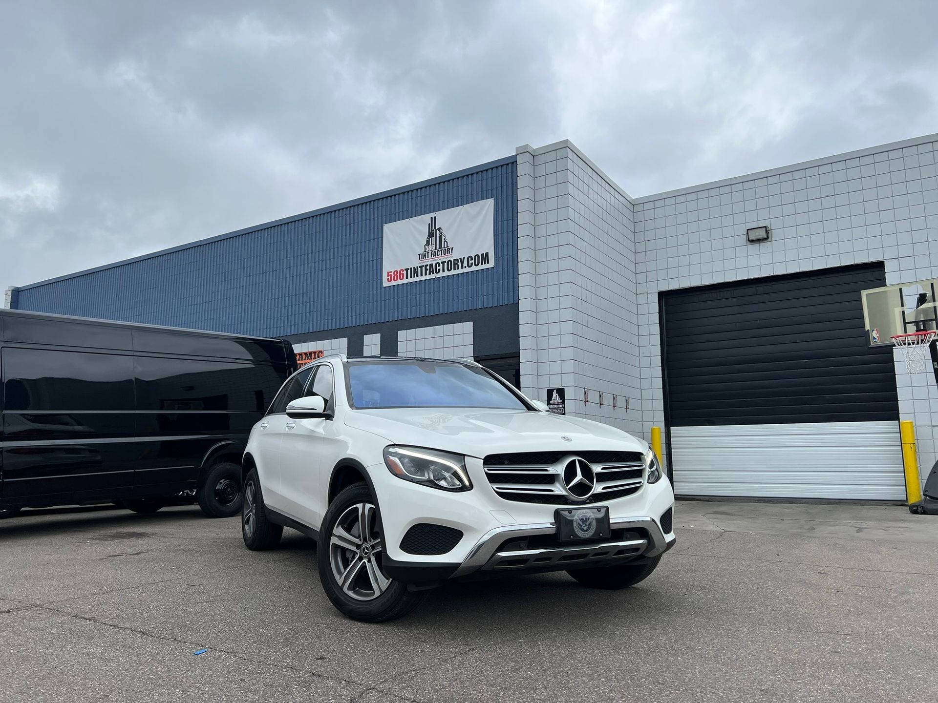 White Mercedes SUV parked outside a gray automotive shop with a black van nearby.