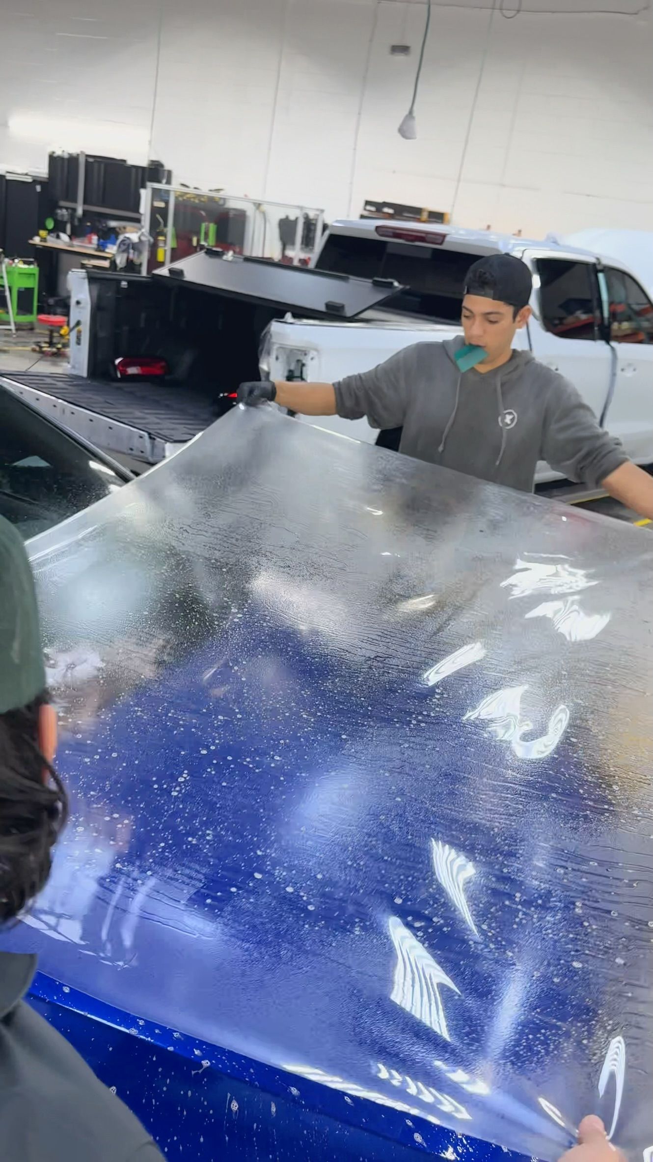 Person in a workshop handling a large transparent sheet over a blue car hood