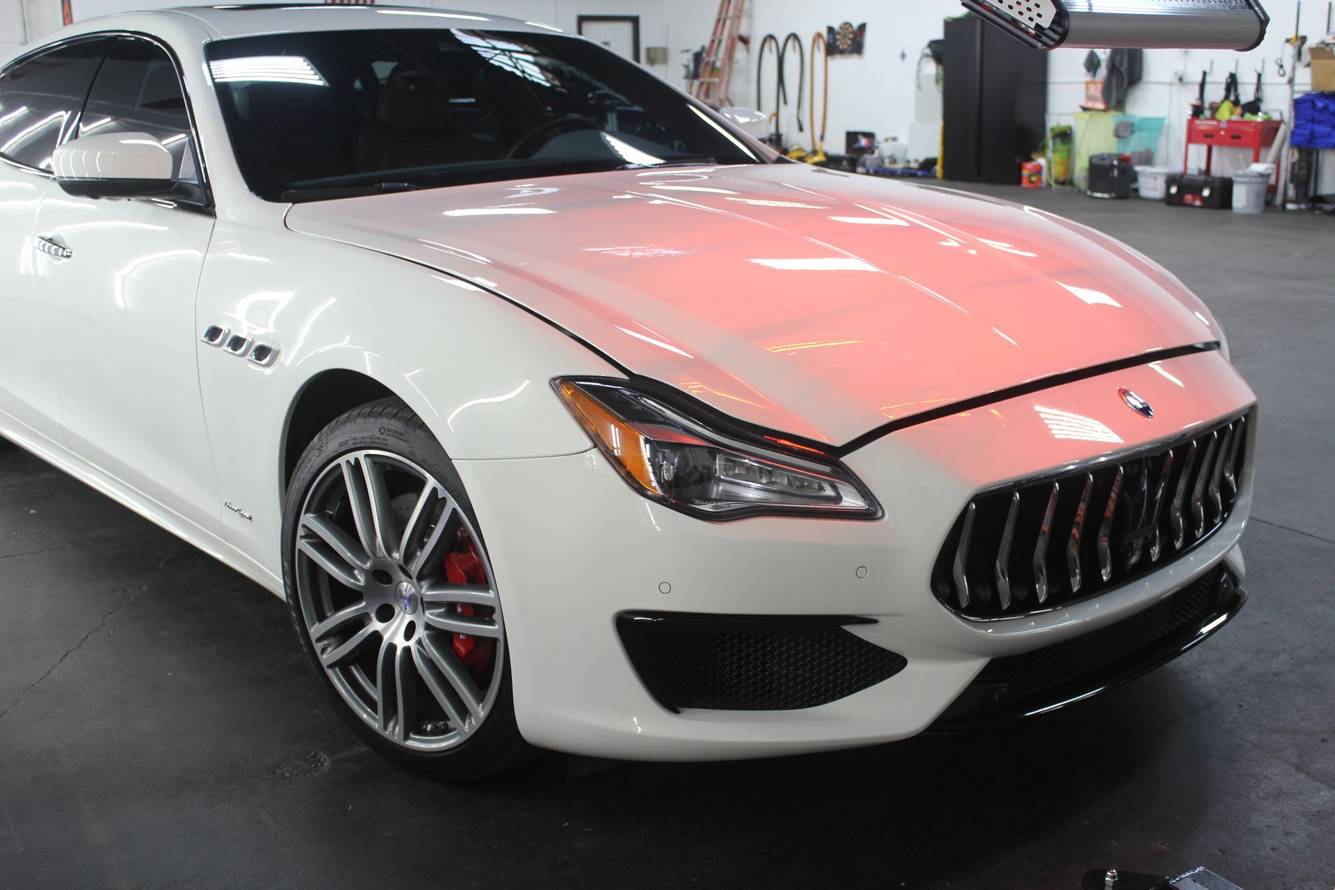 White Maserati sedan in a garage, shown from the front-left with red interior reflections on the hood.