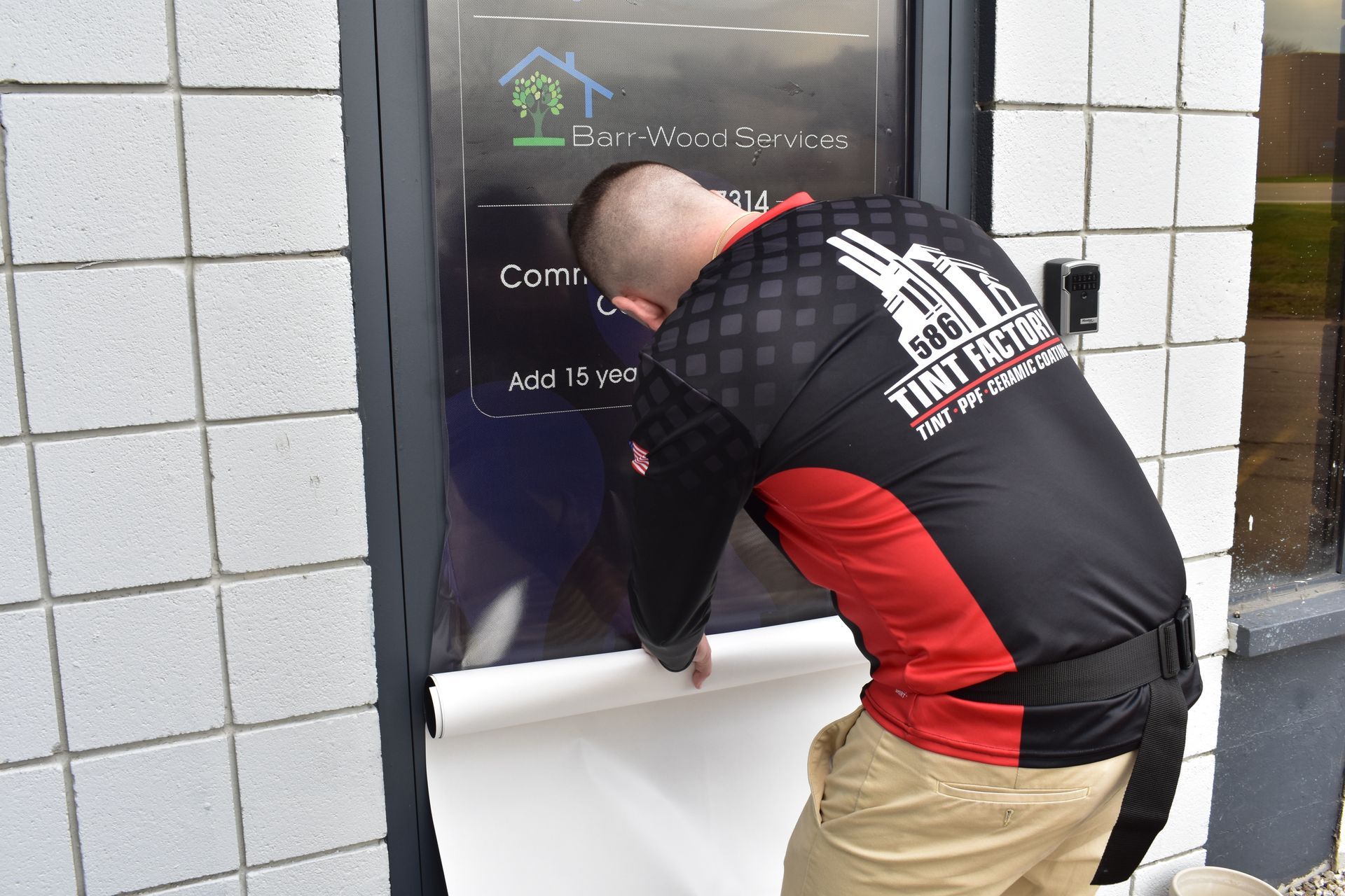 Man in black and red shirt working on a storefront sign beside white tiled wall