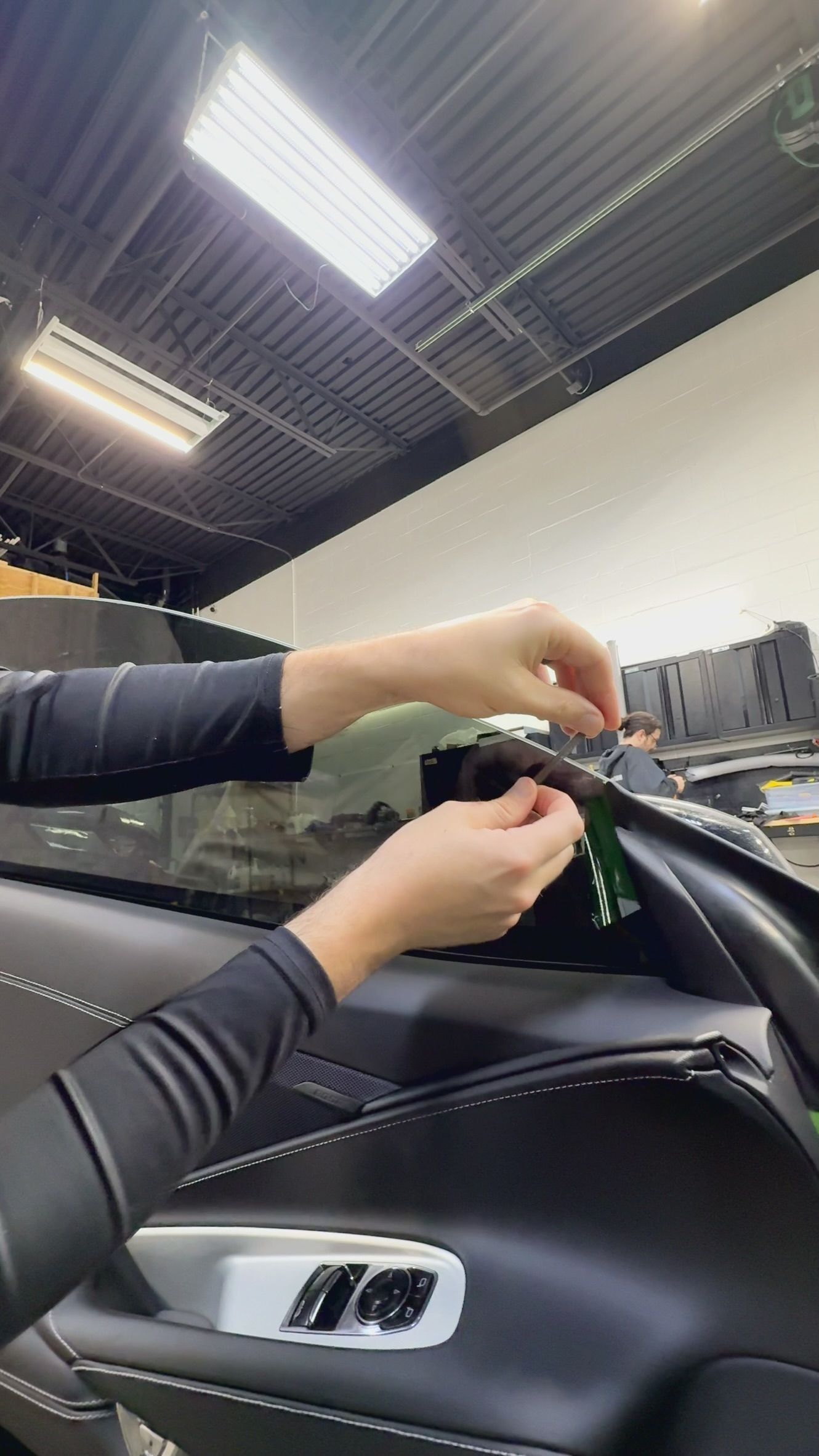 Hands installing a black car part inside a vehicle interior under bright workshop lights