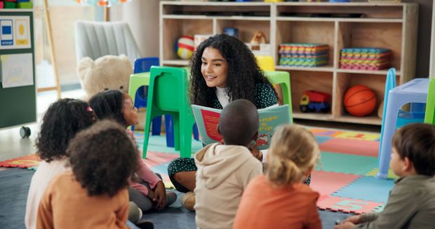 Teacher reading to children seated on a colorful classroom rug, with shelves of toys and books behind them