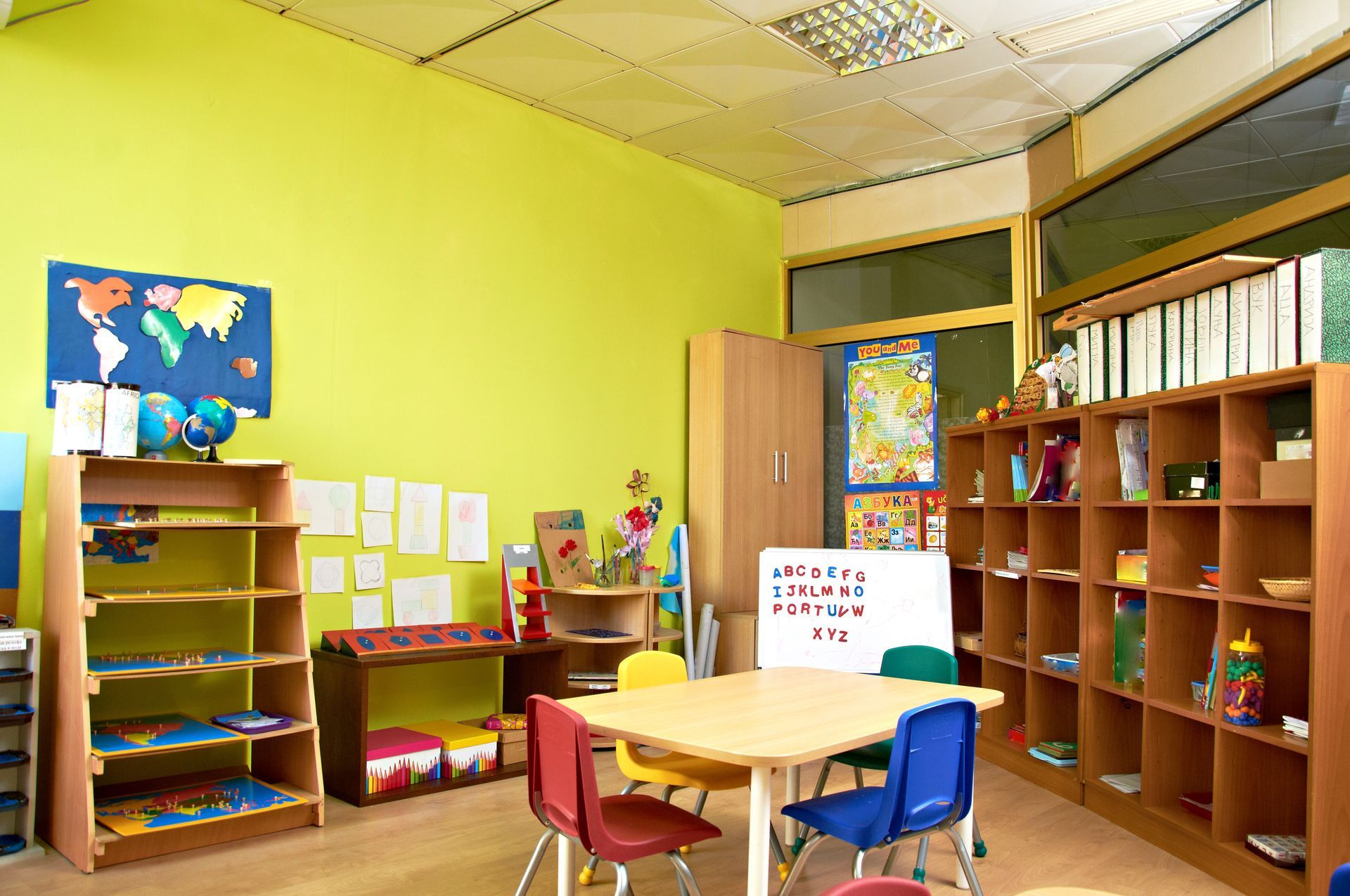Bright classroom with colorful shelves, small tables, chairs, and posters on lime-green walls.