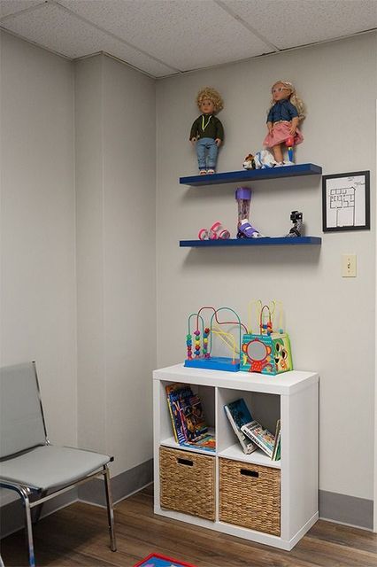 Corner of a therapy/playroom with shelves of dolls, toys, and a white storage cabinet with baskets.