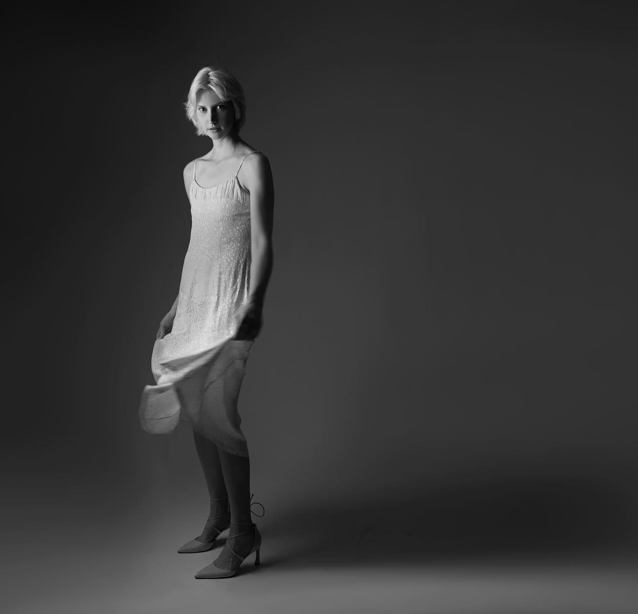 Woman in a white dress and heels, standing and lifting the hem, in a studio with dramatic lighting.