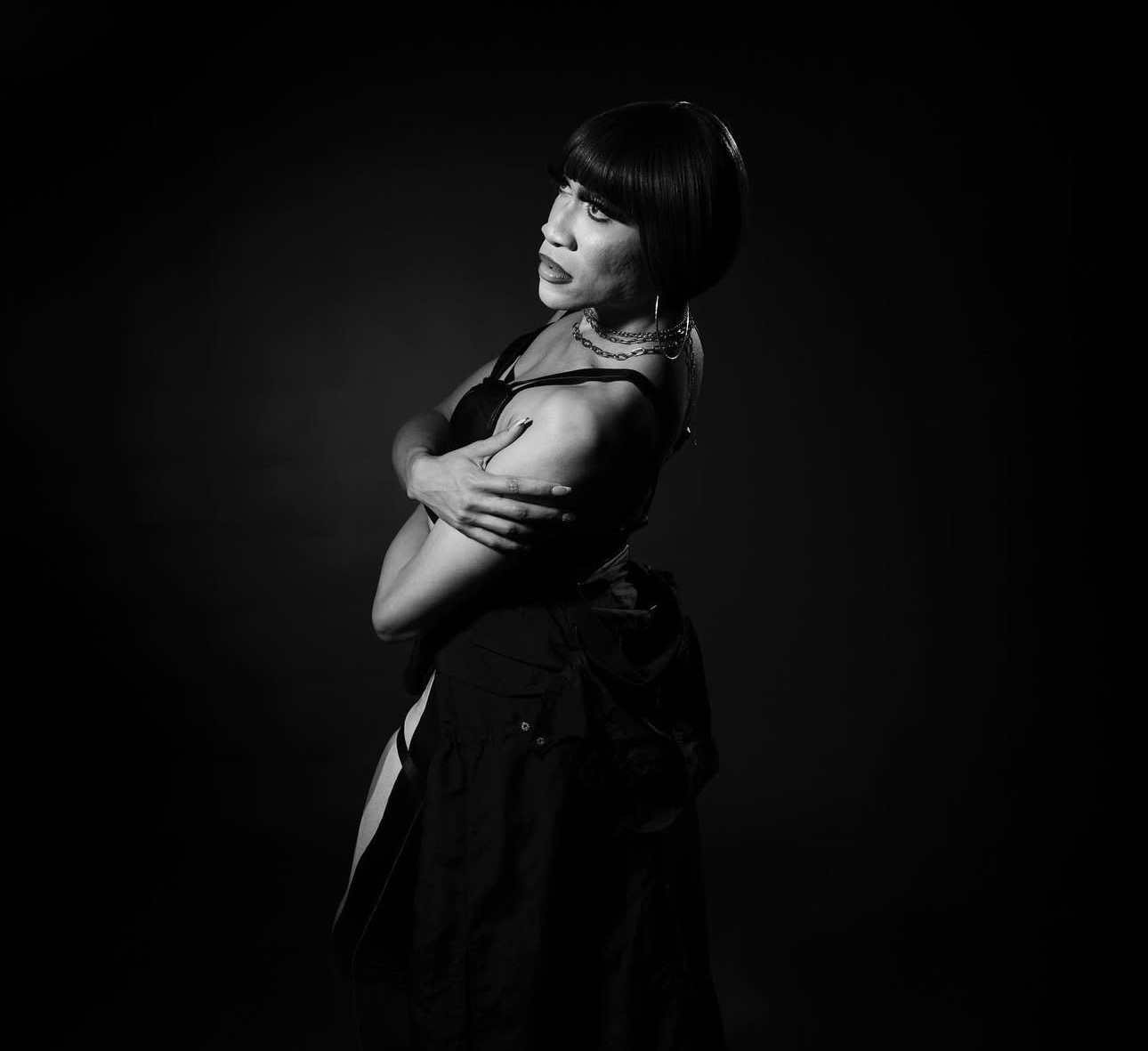 Woman in dark dress, arms crossed, looking over shoulder, against dark background.