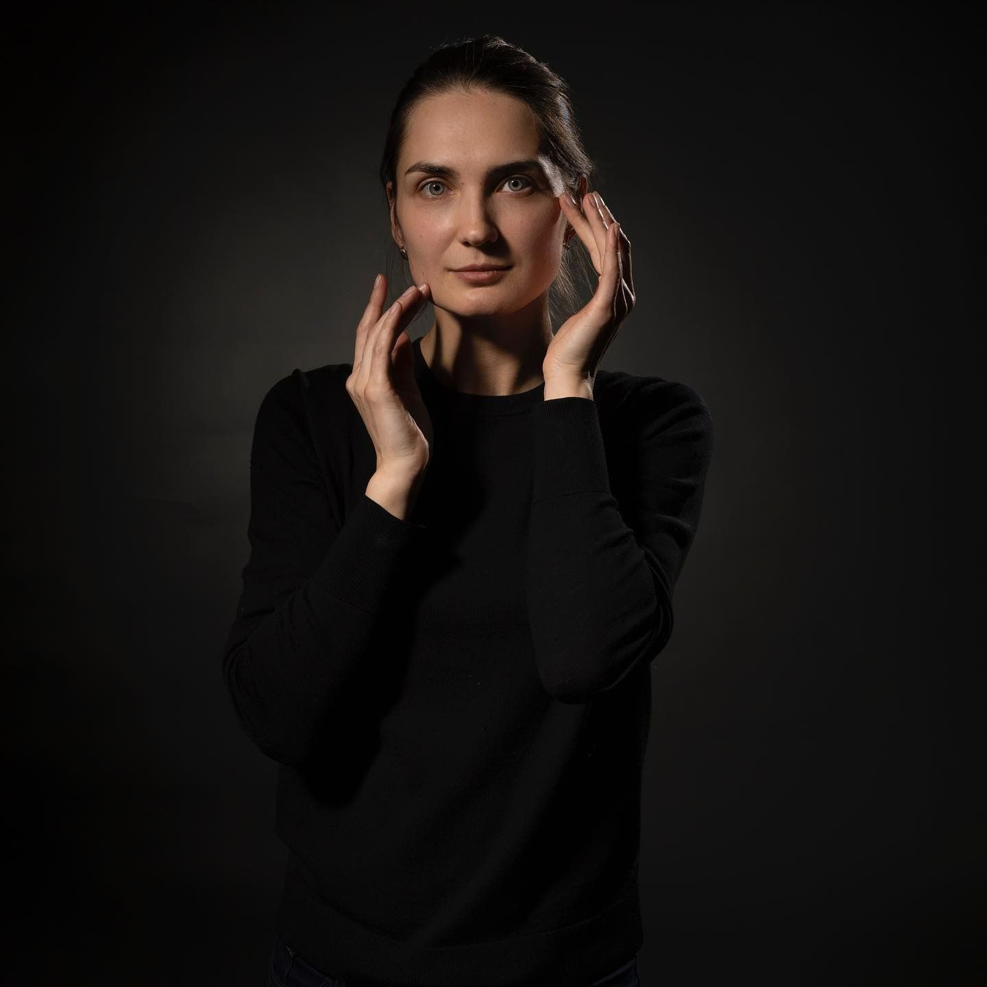 Woman in black sweater with hands near face against dark background.