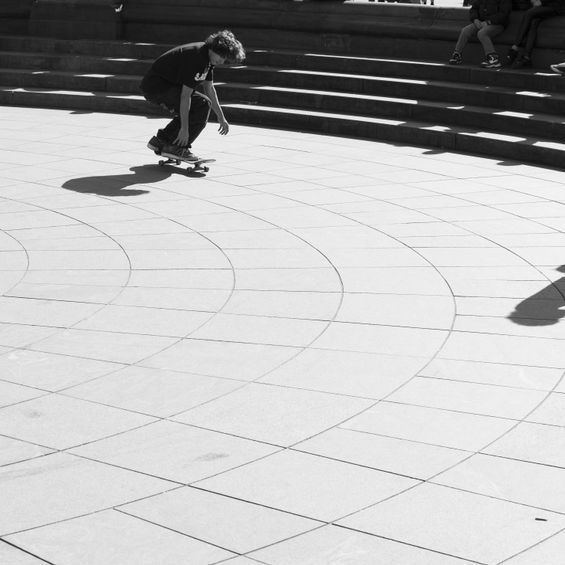 Skateboarder riding on a paved circular area near steps, casting a long shadow.