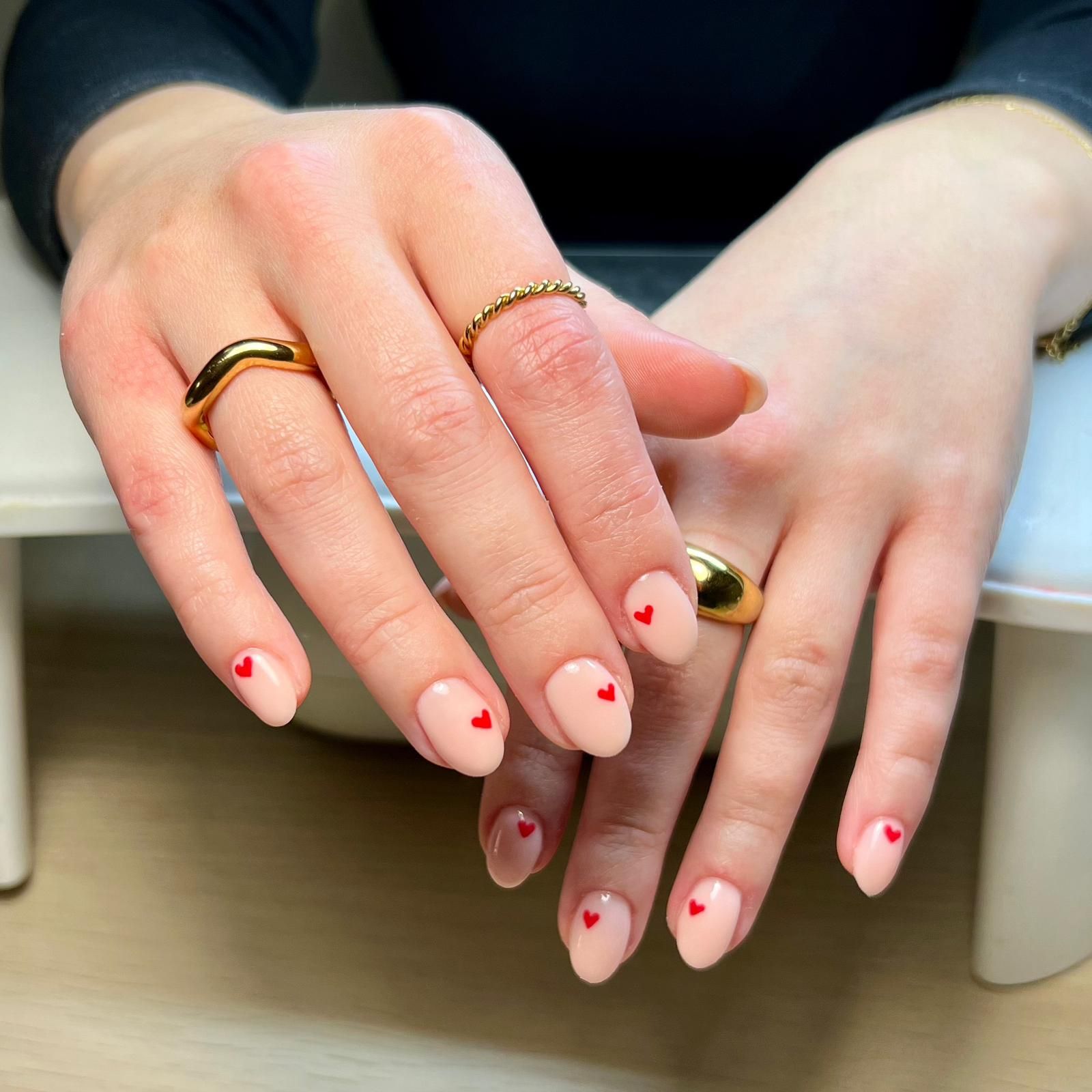 Two hands with pale pink nails and red heart accents, wearing gold rings, resting on a table.