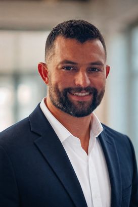 Portrait of Cody smiling in a dark suit and white shirt against a softly blurred indoor background