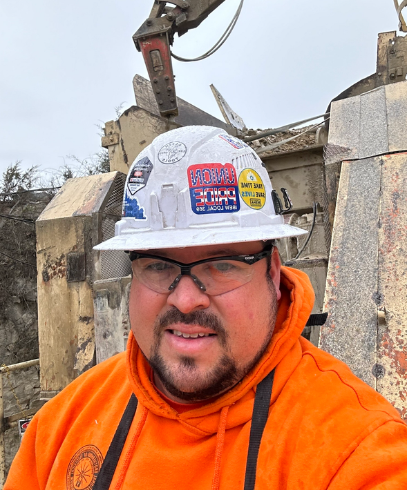 A man wearing an orange hoodie and a hard hat with stickers on it.