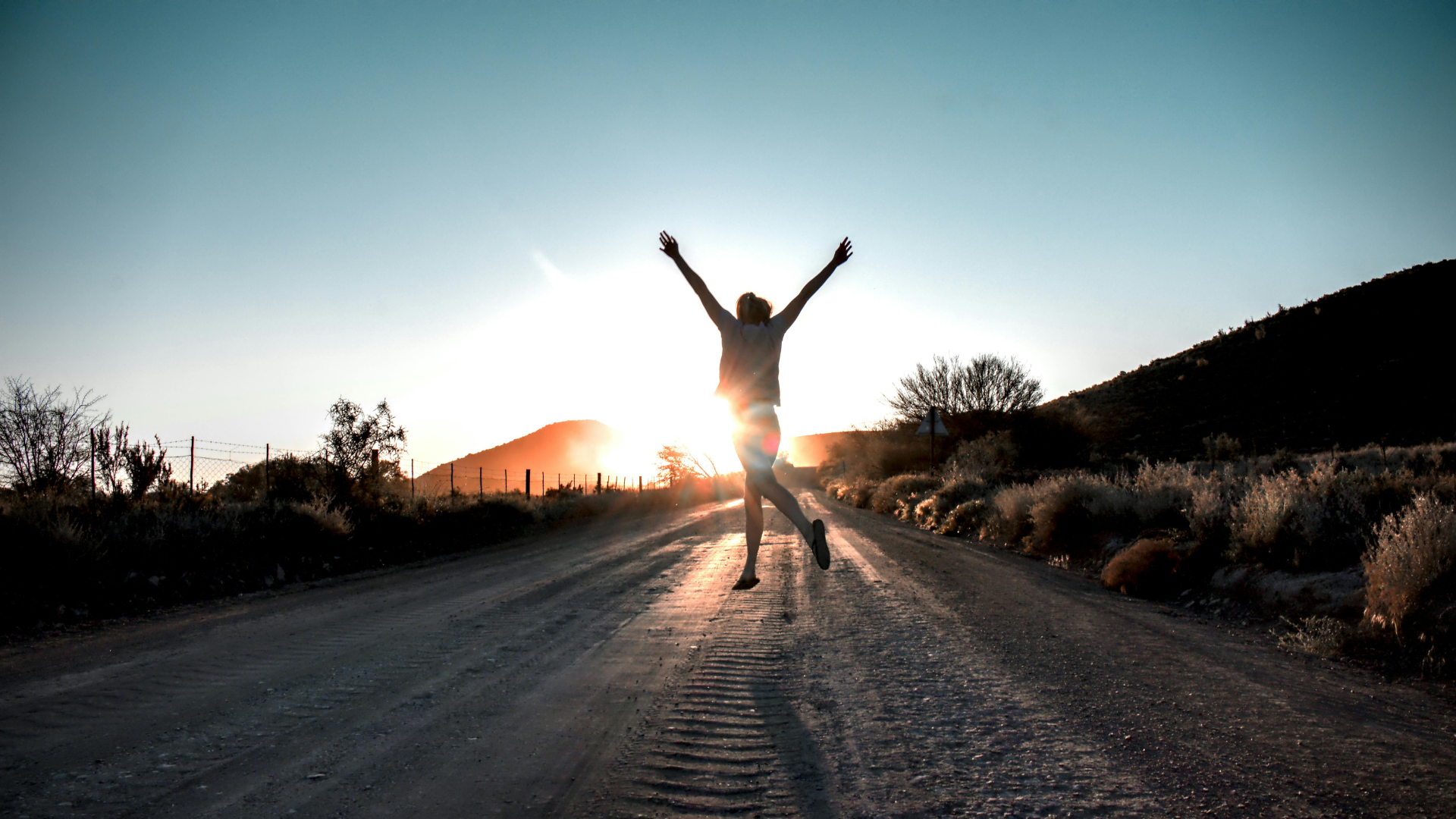 Person jumping on a dirt road, arms raised, sun setting in the background.