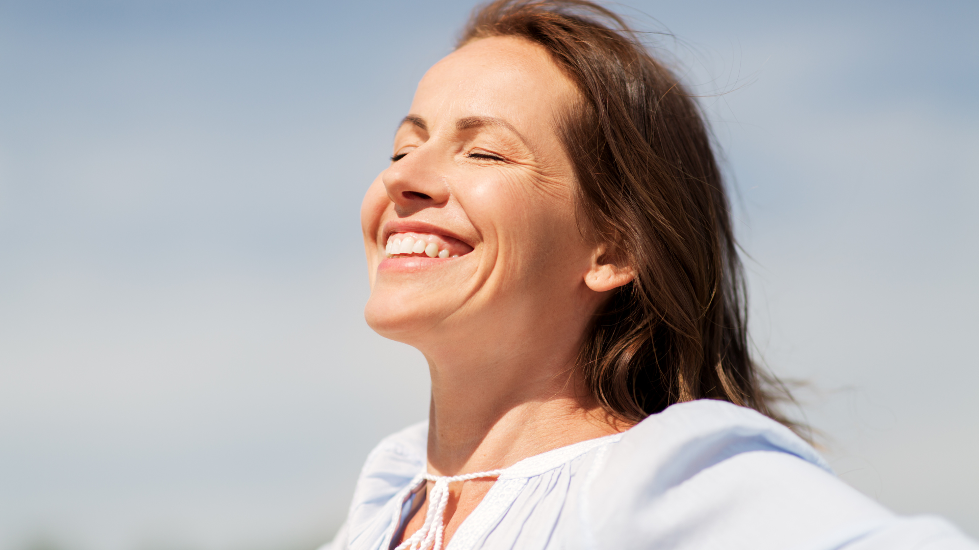 Woman smiles broadly, eyes closed, head tilted back, blue sky background.