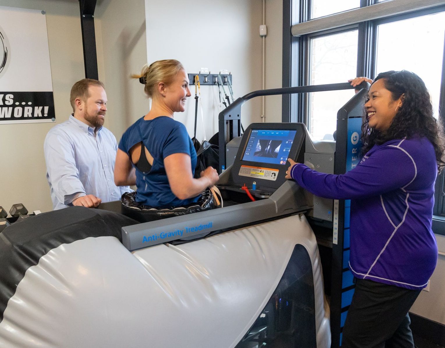 A person in a medical clinic walks on an AlterG anti-gravity treadmill while two staff members monitor the display.