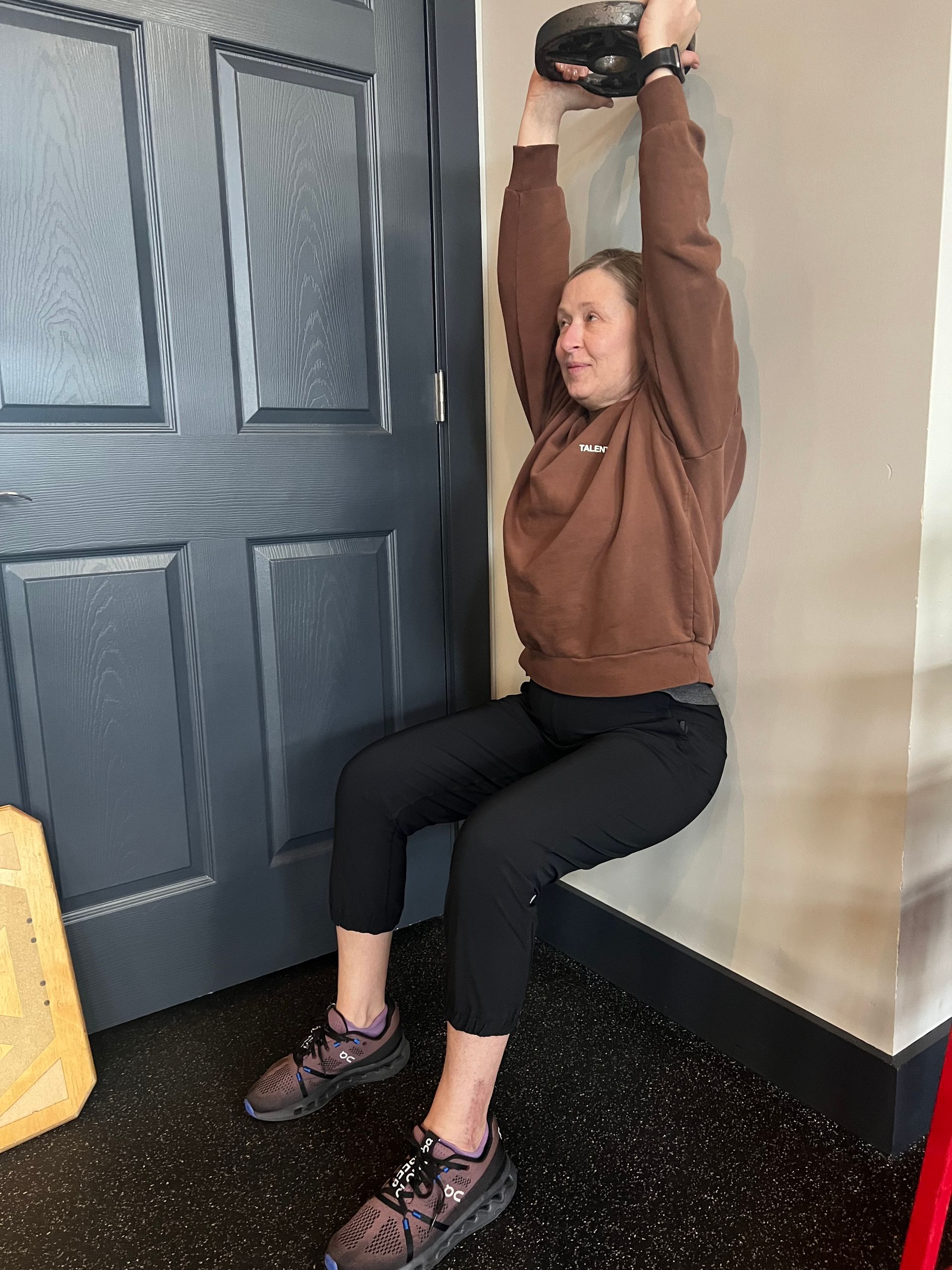 A woman is squatting against a wall while holding a kettlebell.