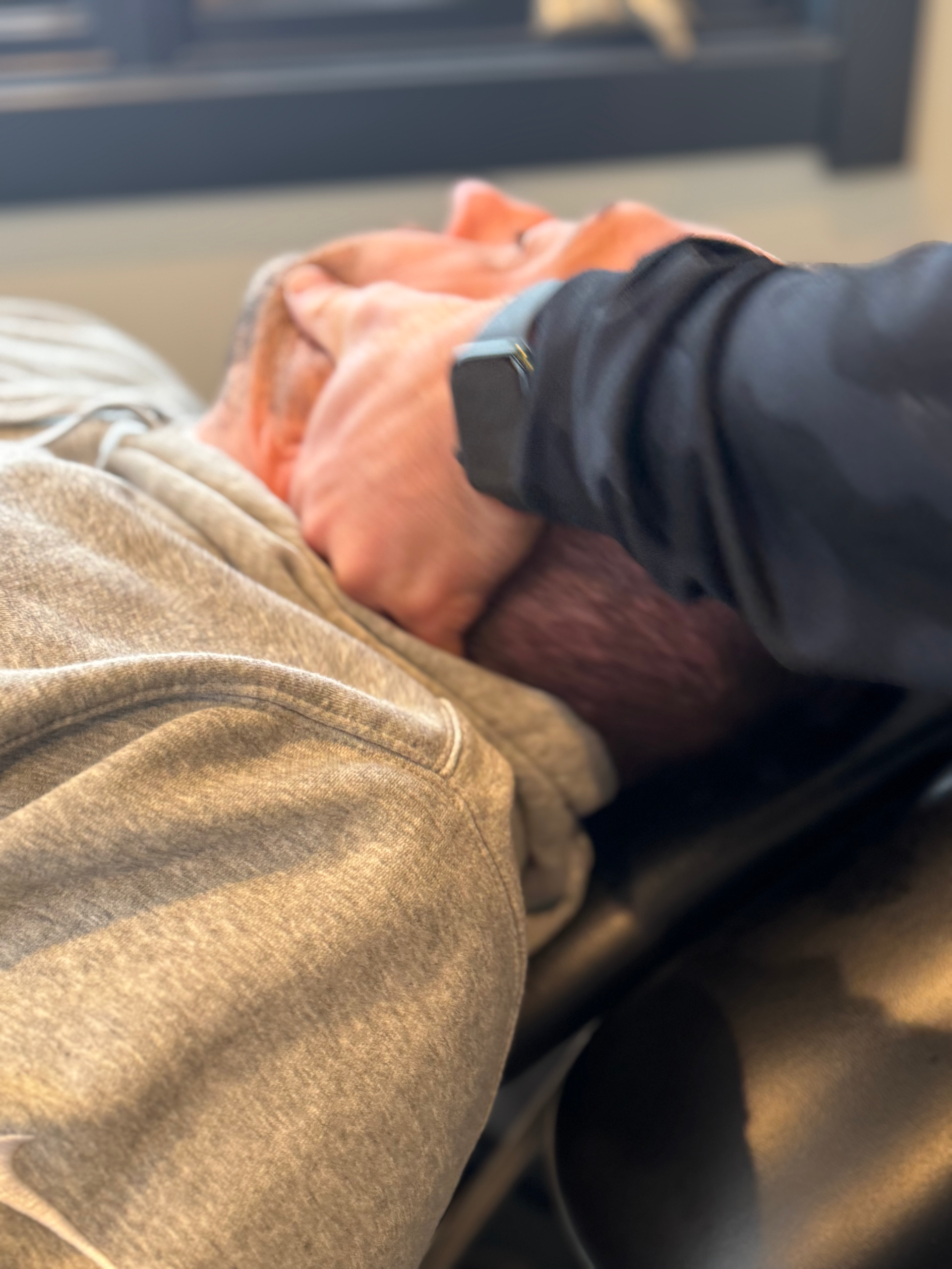 Man reclining on a chair, face covered by a hand, wearing a gray hoodie in a waiting area